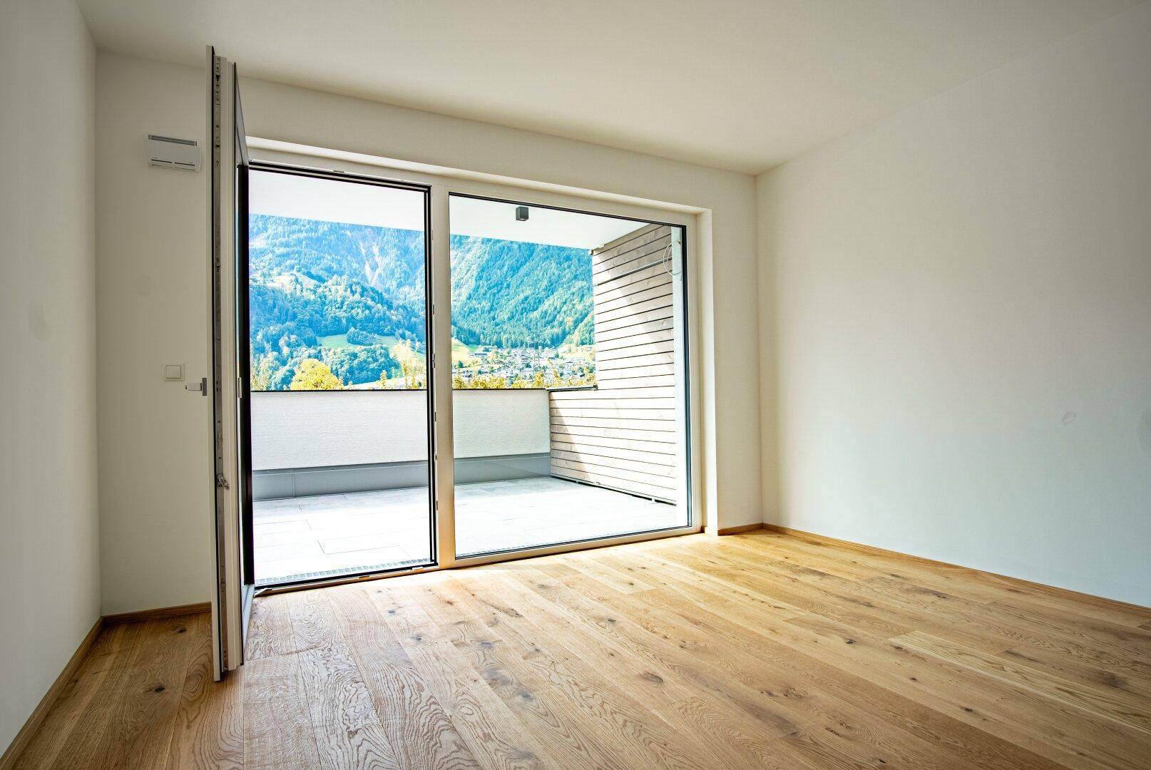 Heller, leerer Raum mit Holzboden und großen Fenstern, die Zugang zu einer Terrasse mit Bergblick bieten.