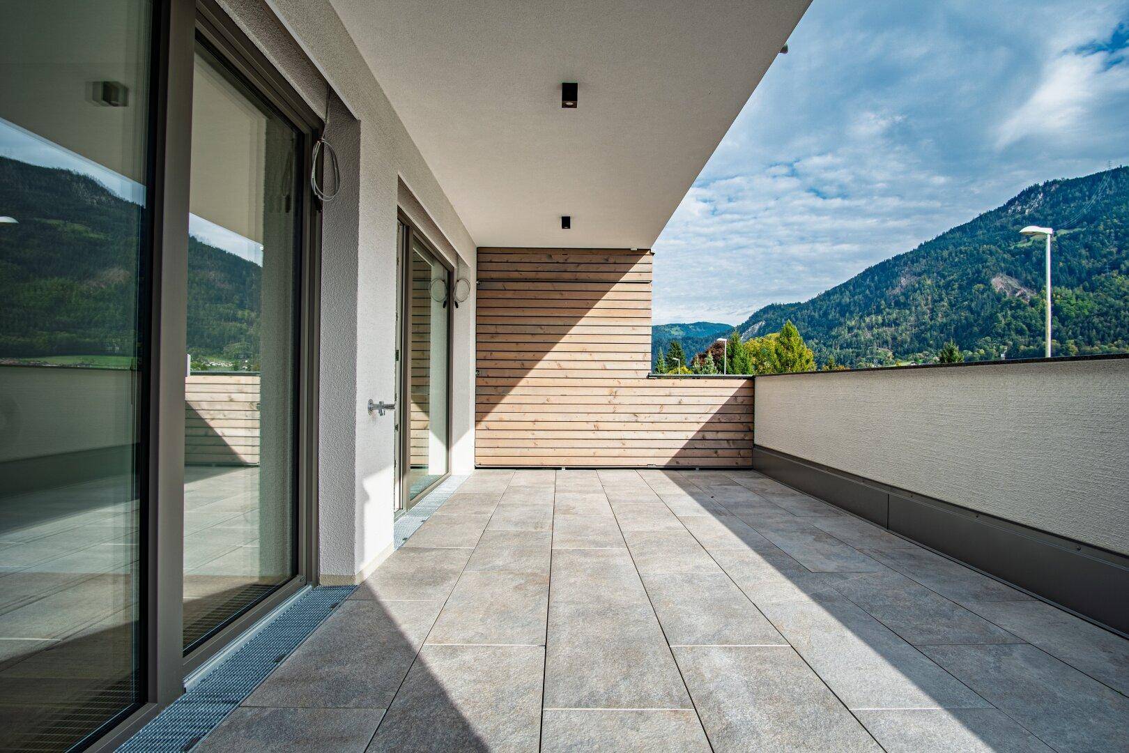 Sonnige Außenterrasse mit grauen Fliesen und Holzverkleidung, die einen weiten Bergblick bietet.