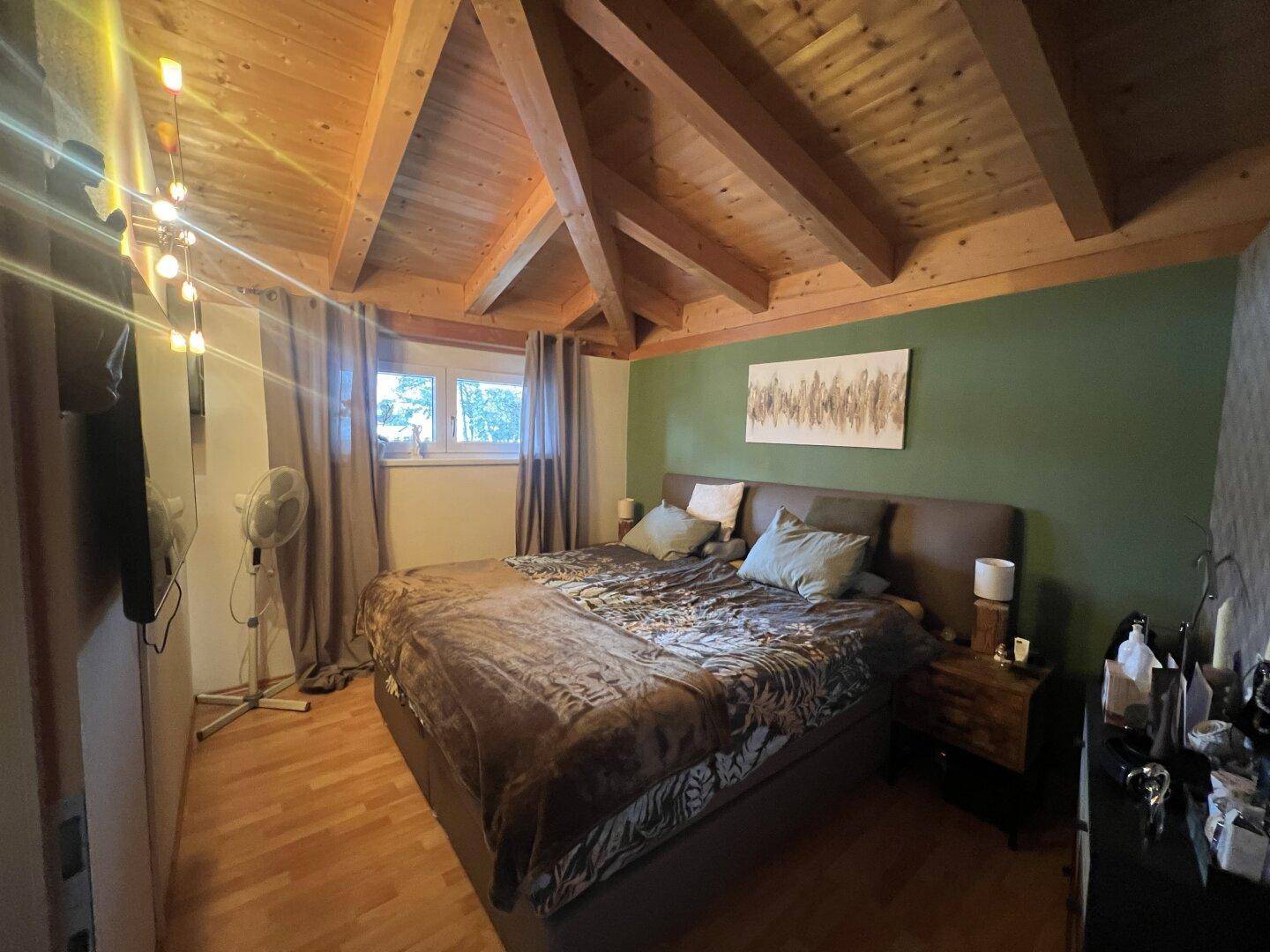Schlafzimmer mit Holzbalkendecke, grüner Akzentwand und Parkettboden im Dachgeschoss.