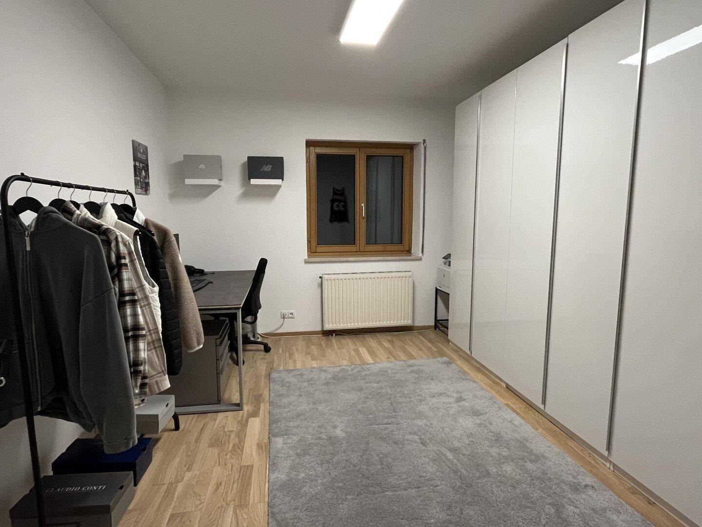 Schlafzimmer mit Holzfußboden, großem Kleiderschrank und Schreibtisch am Fenster.