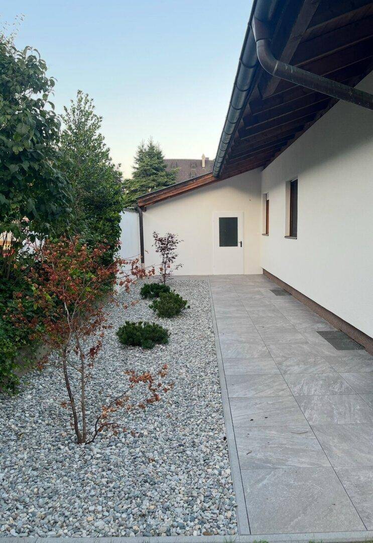 Seitlicher Gartenweg mit Kiesbeet, jungen Bäumen und gepflasterter Fläche am Haus.
