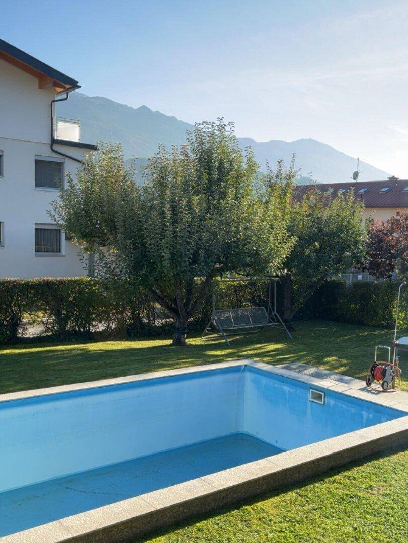 Gartenbereich mit Pool, Rasenfläche, Bäumen und Schaukel vor Bergkulisse.