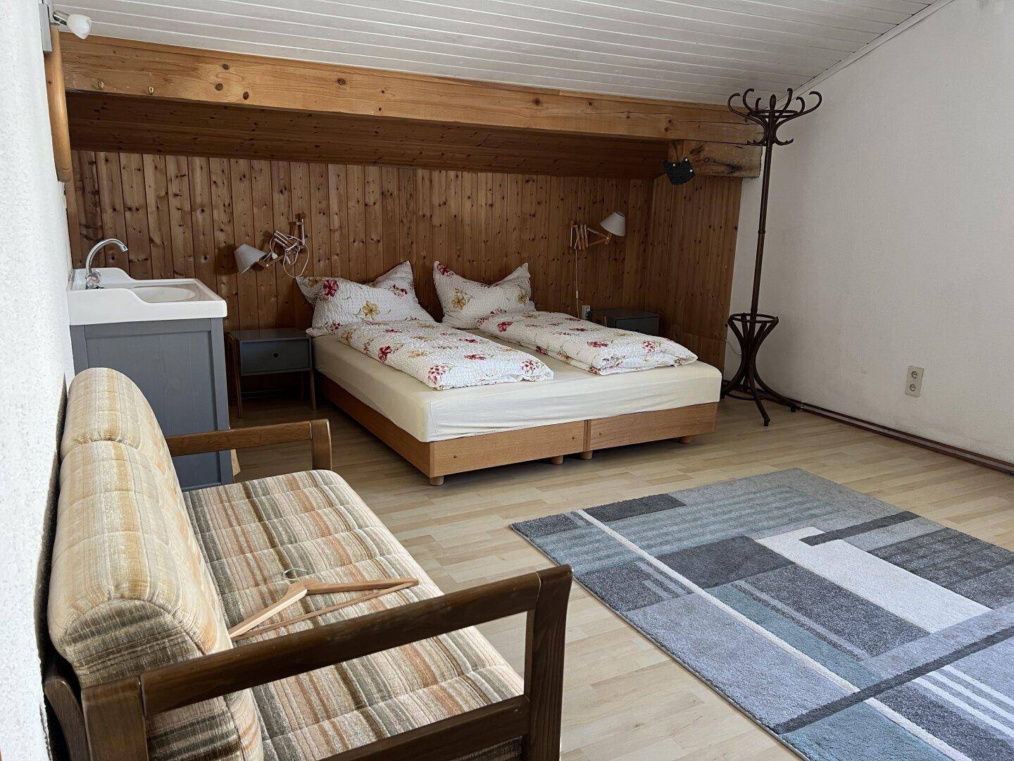 Schlafzimmer mit Holzwänden, Doppelbett und kleinem Waschbecken in der Ecke.