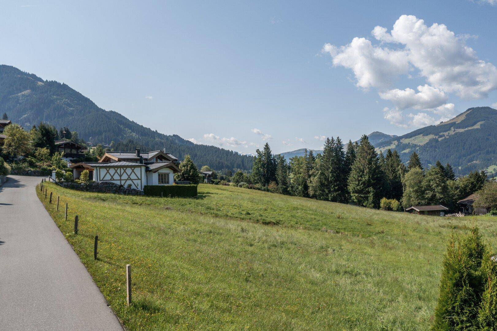 Weitläufige grüne Wiesenfläche mit Zufahrtsstraße und Blick auf die umliegende Berglandschaft.