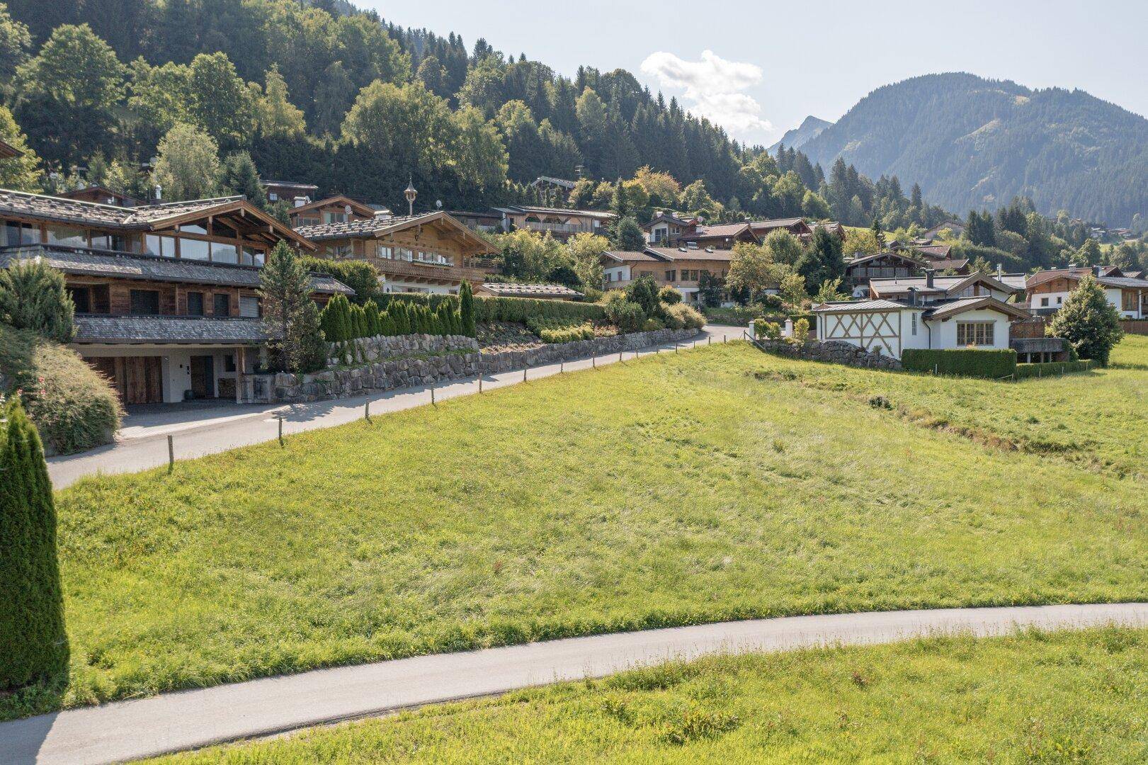 Erhöhte Ansicht eines großen Wiesengrundstücks mit Zufahrtsweg und traditionellen Häusern im Hintergrund.