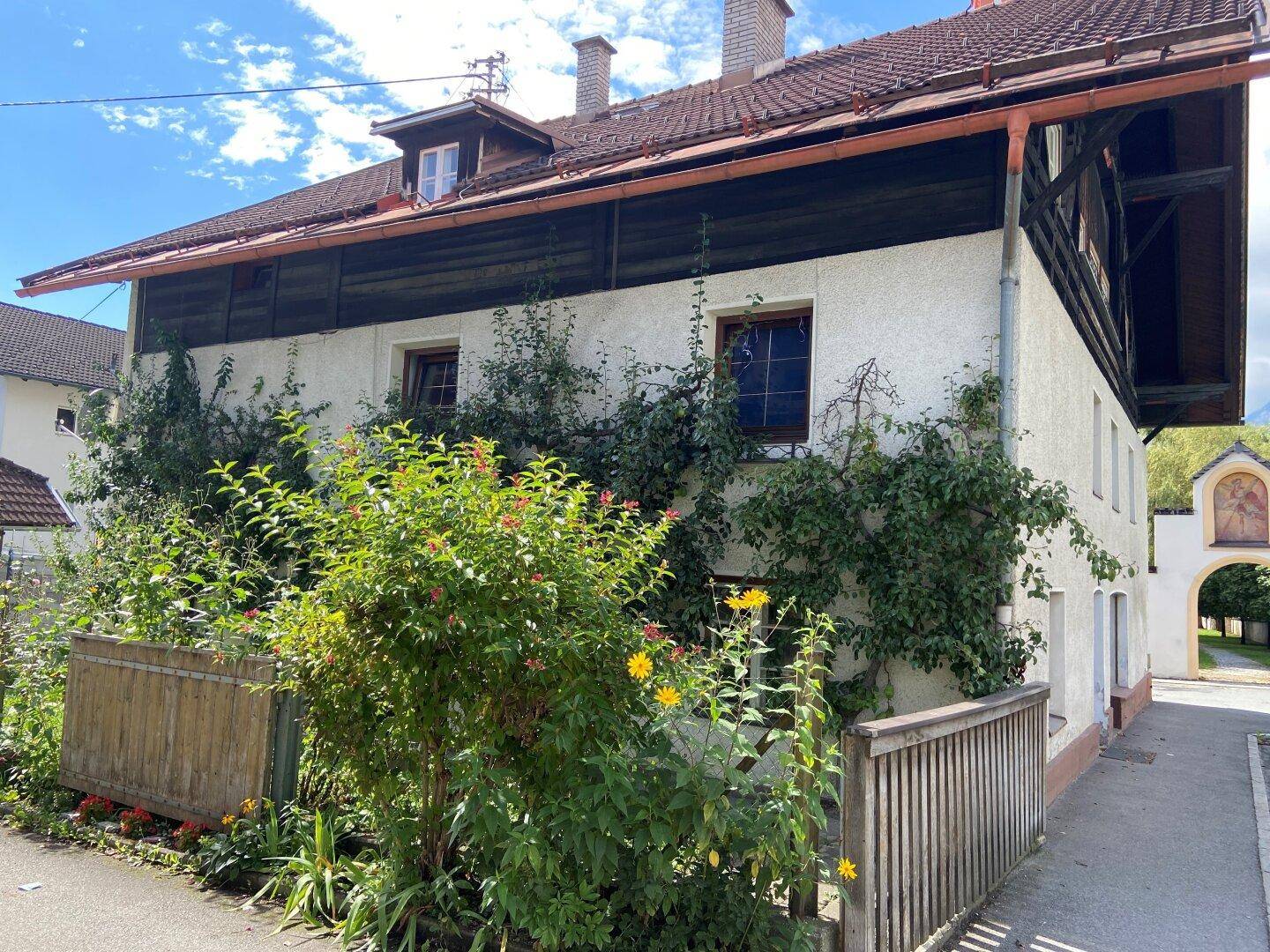 Seitenansicht eines älteren Wohnhauses mit üppiger Begrünung und traditionellem Dach.