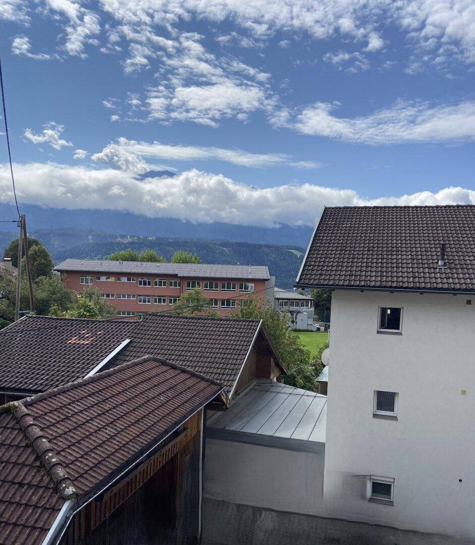 Weitblick über Dächer und grüne Hügel bis zu den majestätischen Bergen unter blauem Himmel.
