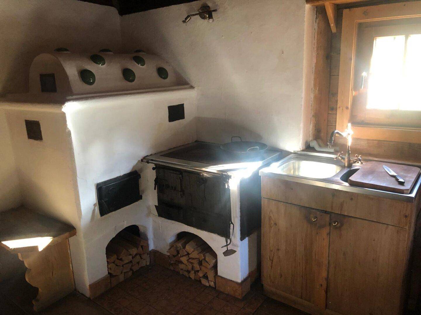Rustikale Küche mit traditionellem Kachelofen, Holzschränken, Spülbecken und natürlichem Lichteinfall durch das Fenster.