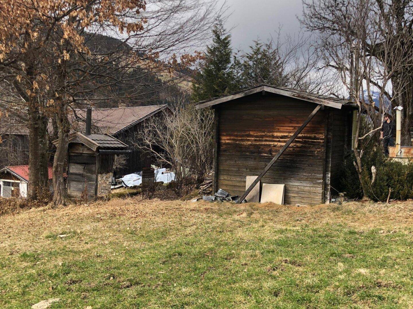 Zwei kleine, rustikale Holzschuppen auf einem ländlichen Grundstück, umgeben von Bäumen und Gras.