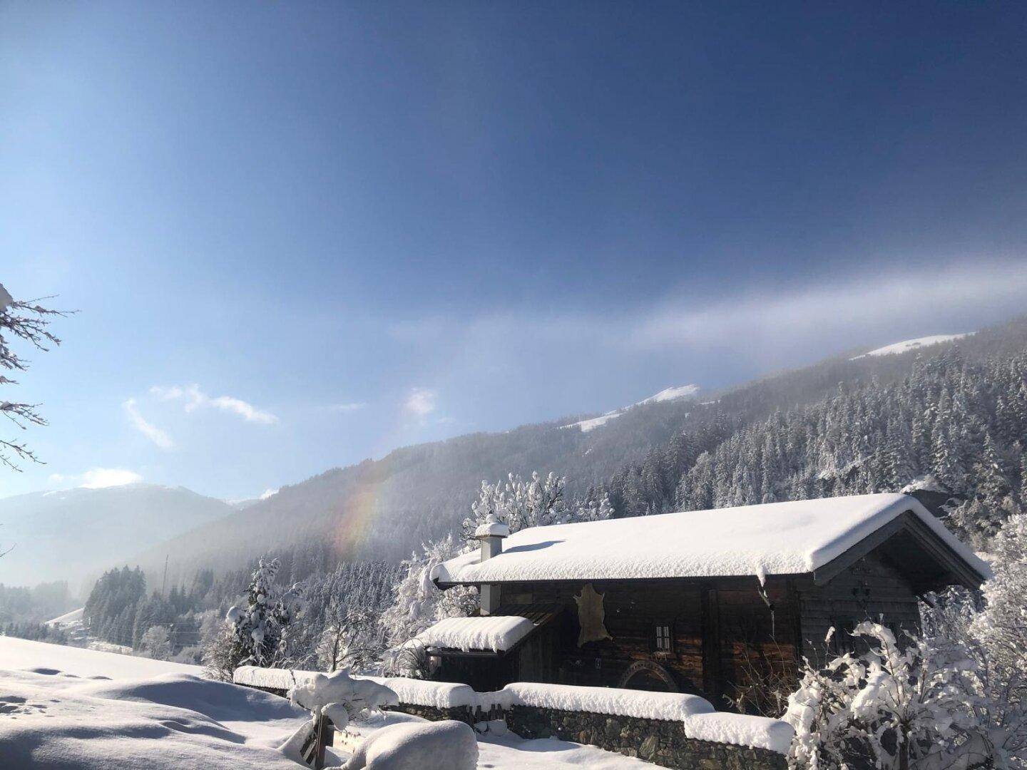 Verschneites Holzhaus in einer idyllischen Winterlandschaft mit majestätischen Bergen im Hintergrund und klarem Himmel.