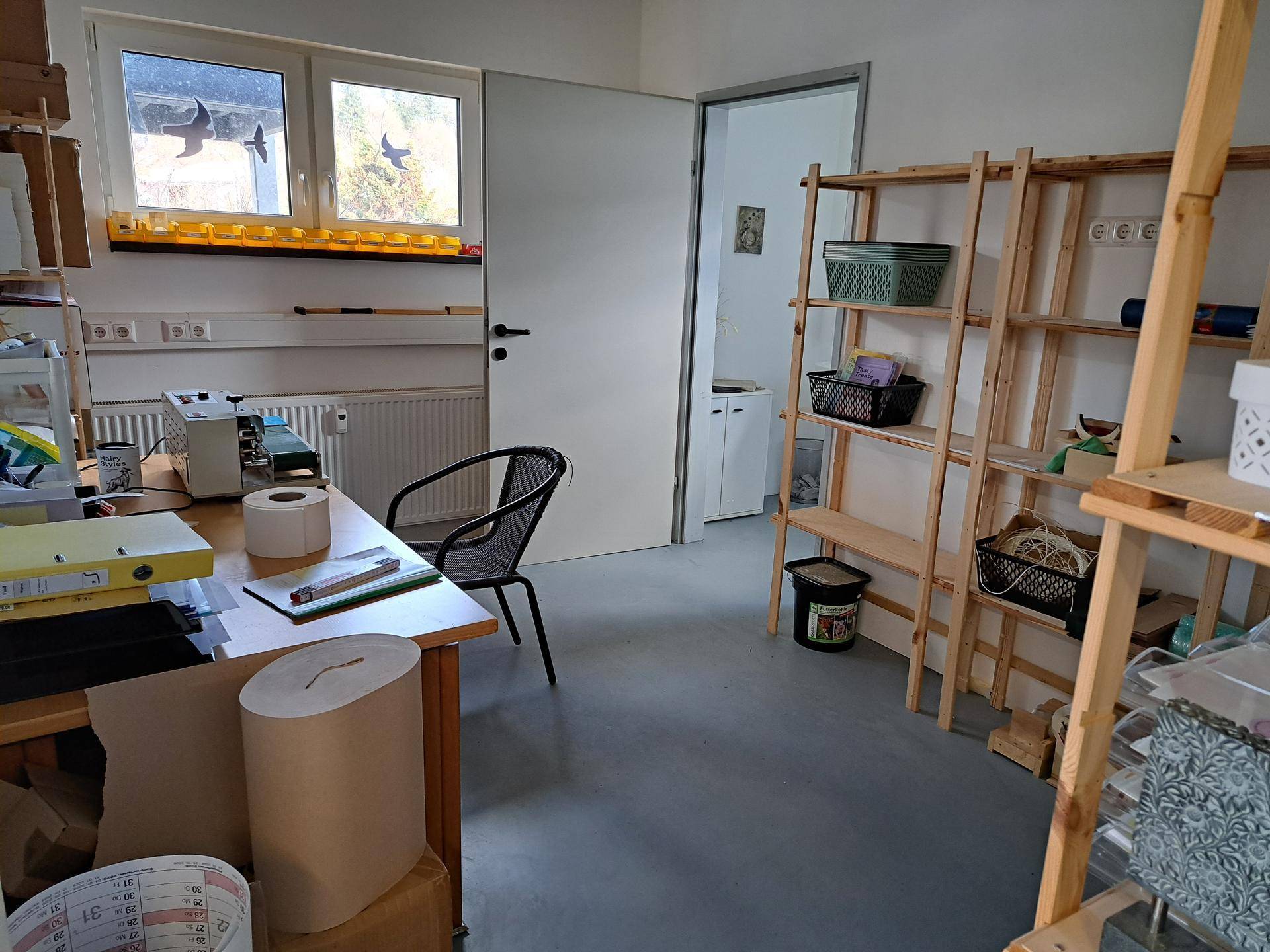 Helles Büro oder Werkstatt mit Schreibtisch, Holzregalen und Fenstern mit Blick ins Grüne.