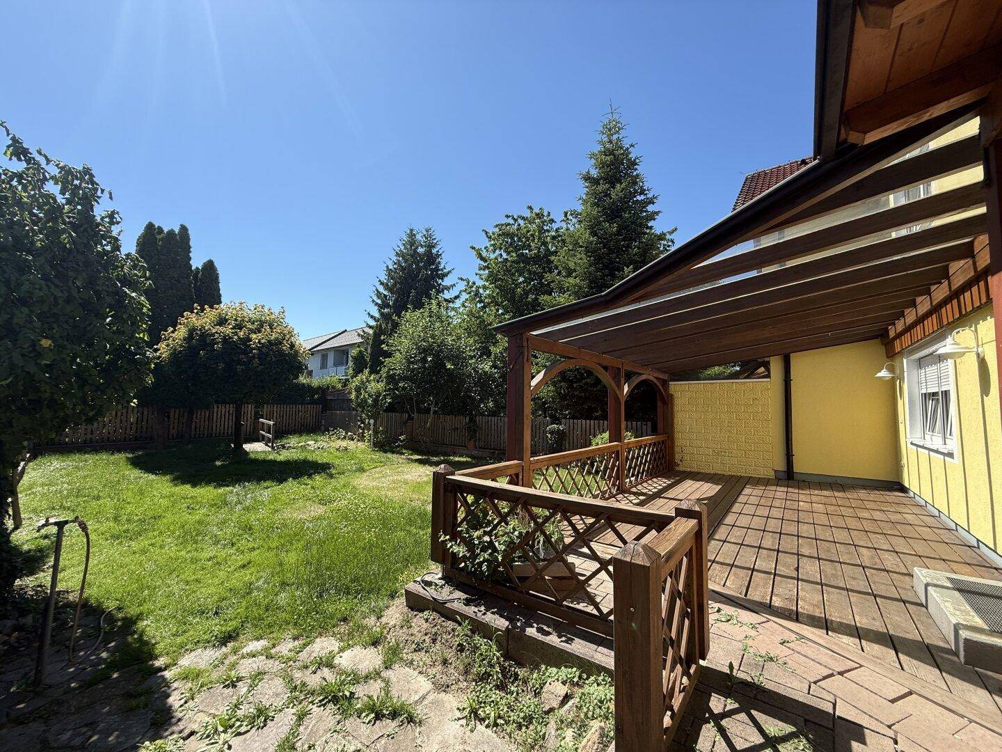 Sonniger Garten mit Rasenfläche und angrenzender Holzterrasse, umgeben von Bäumen und Sträuchern.