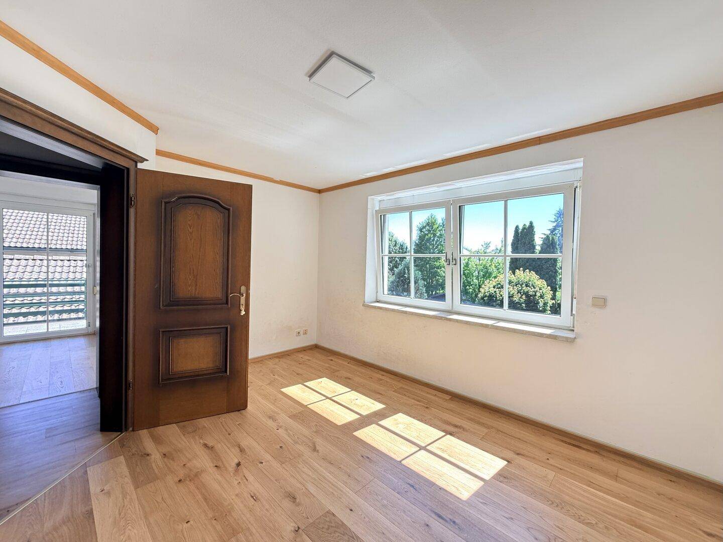 Geräumiges Schlafzimmer mit hellem Holzboden und großem Fenster mit Blick ins Grüne.