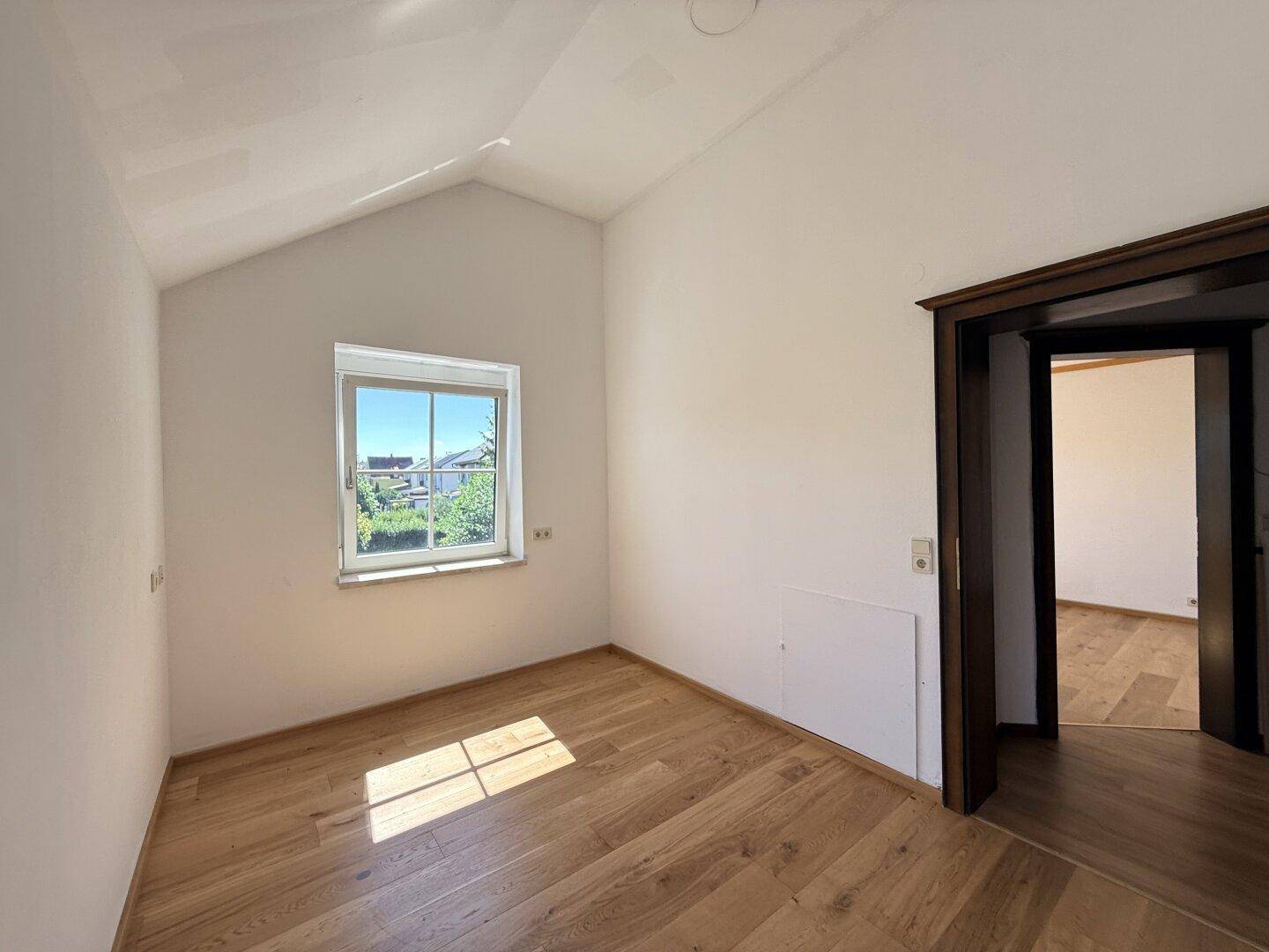 Gemütliches Zimmer im Dachgeschoss mit schräger Decke, Holzboden und Fenster mit Ausblick.