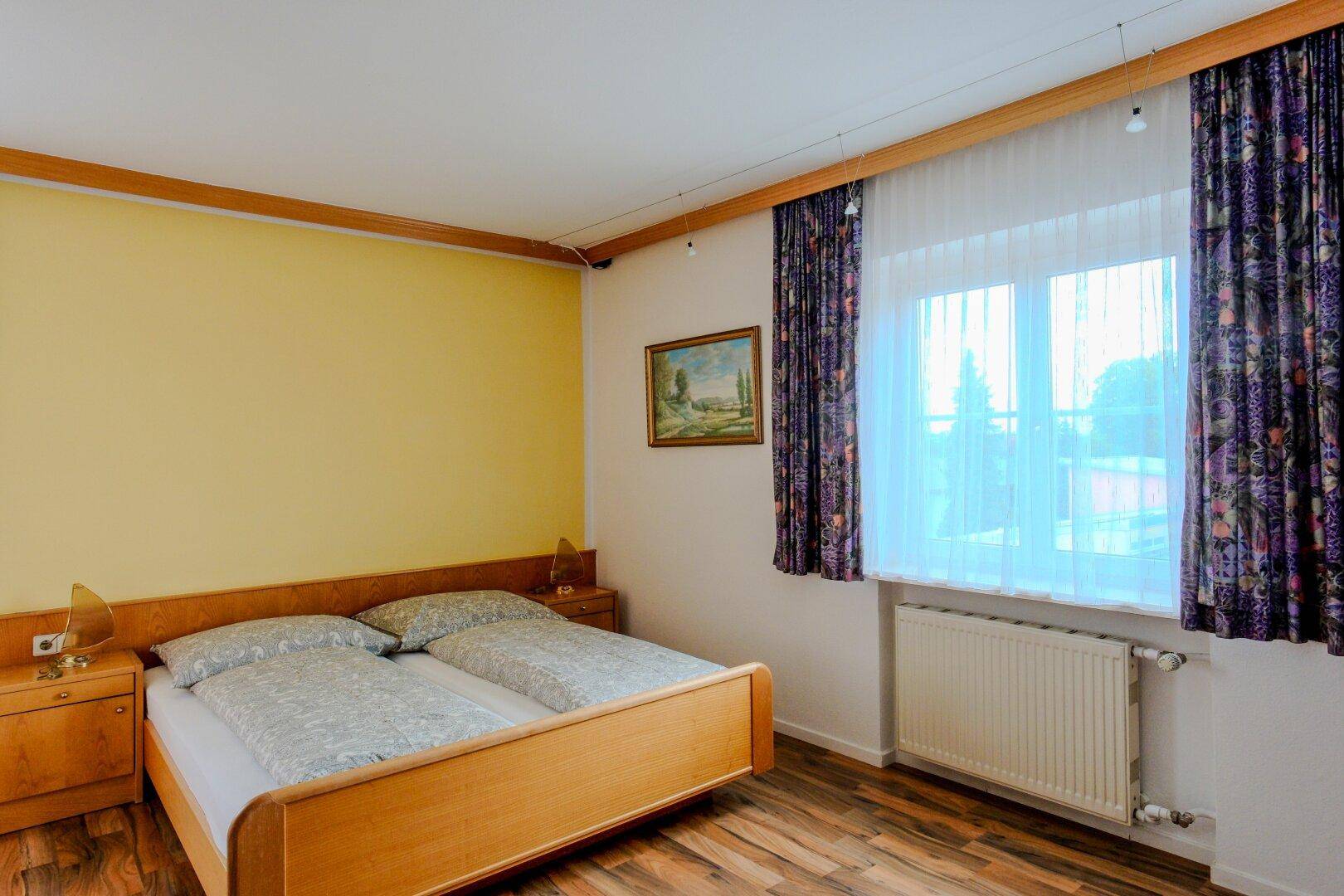 Helles Schlafzimmer mit Doppelbett, Nachttischen und gemusterten Vorhängen an den Fenstern.