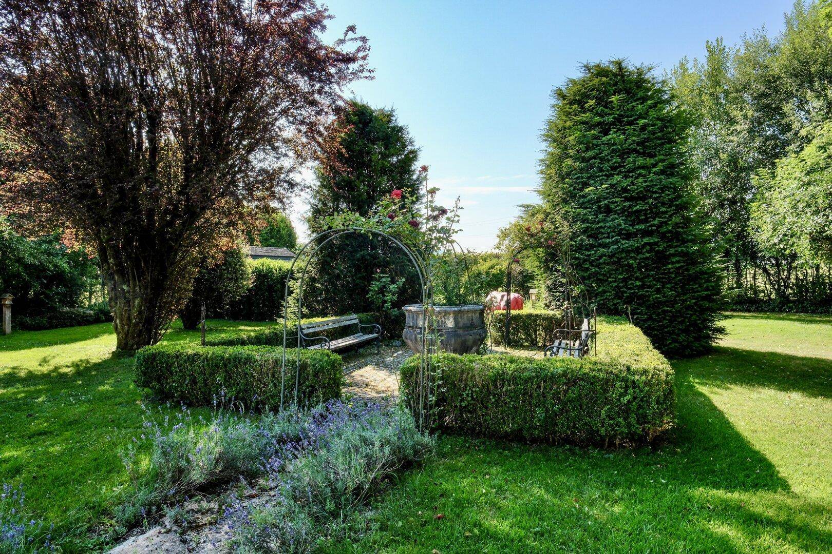 Idyllischer Gartenbereich mit gepflegten Hecken, Rosenbögen und einer Steinbank unter hohen Bäumen.