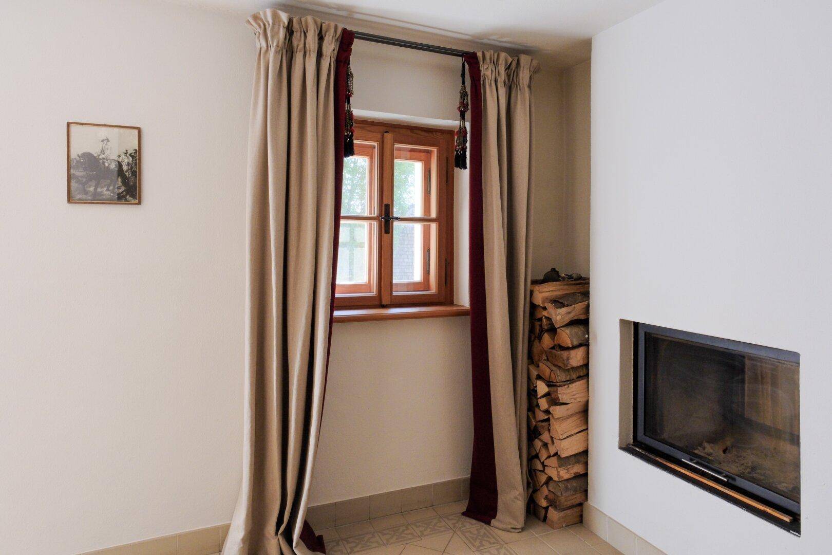 Heller Raum mit integriertem Kamin, Holzstapel und Fenster mit schweren Vorhängen.