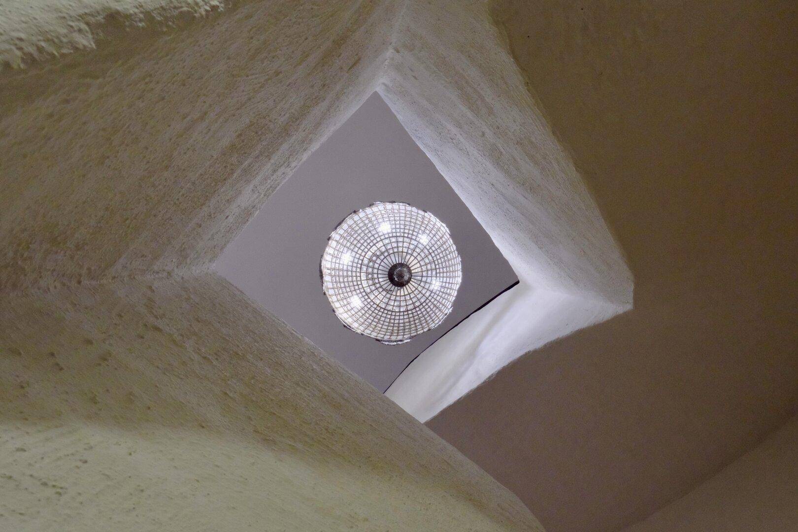 Blick von unten auf einen kunstvollen Kronleuchter, der in einem hohen, quadratischen Lichtschacht hängt.