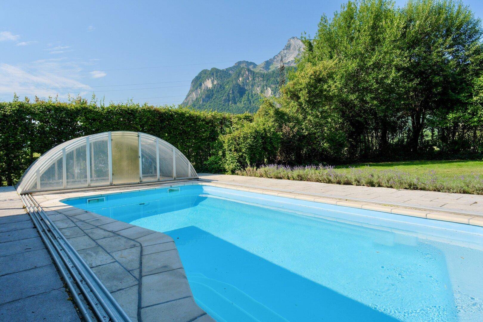 Außenpool mit Abdeckung, umgeben von einer grünen Hecke und Bäumen, mit majestätischem Bergblick.