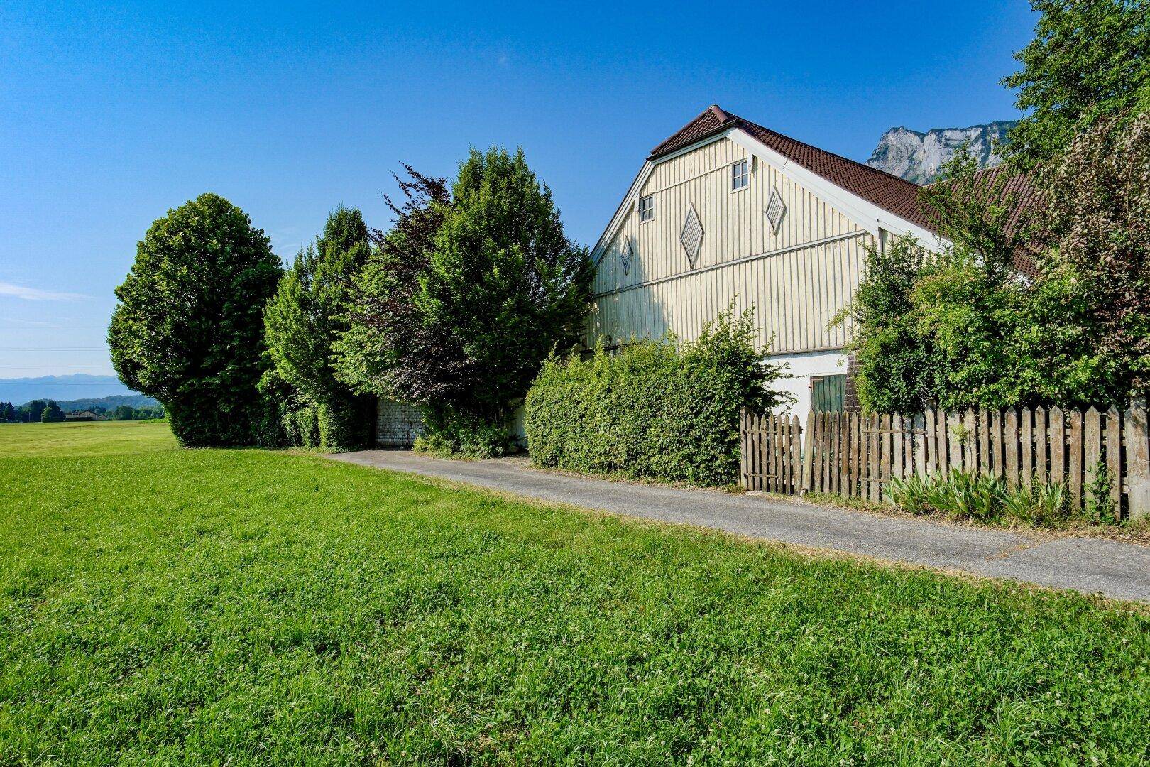 Ländliches Haus mit Holzfassade und Zaun, umgeben von grünen Feldern und einer beeindruckenden Bergkulisse.