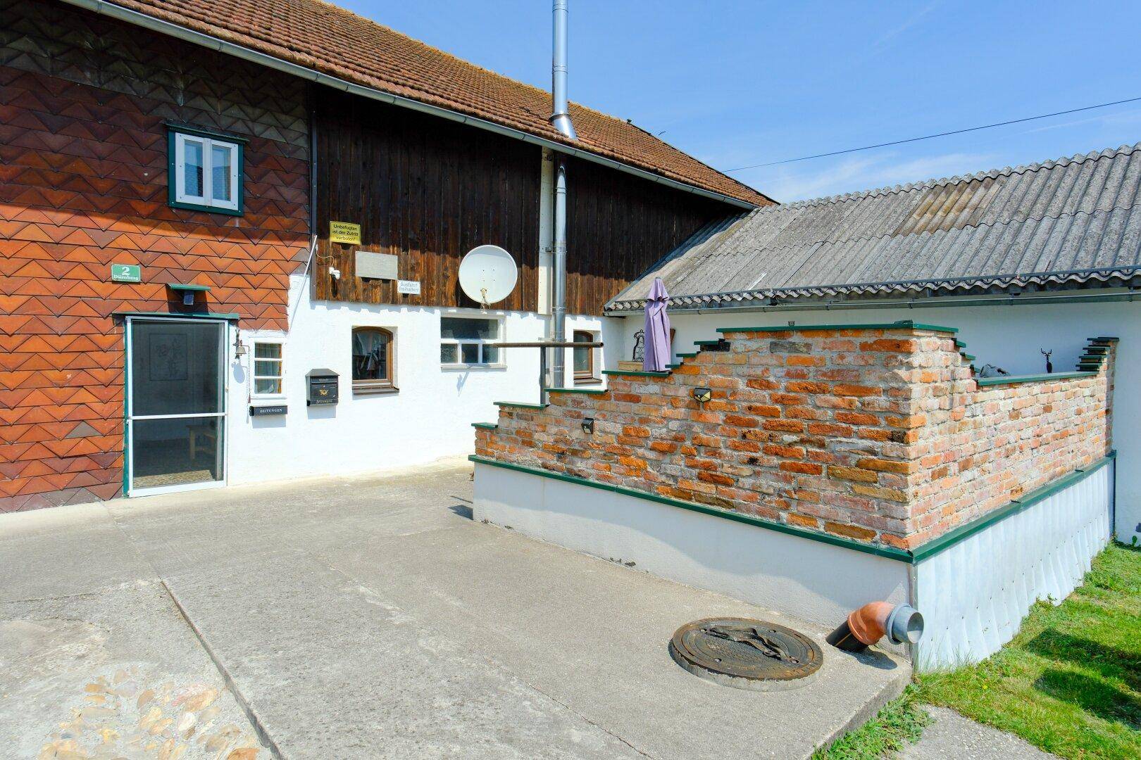 Eingangsbereich eines Bauernhauses mit Ziegelmauer und Zugang zu einem Nebengebäude.