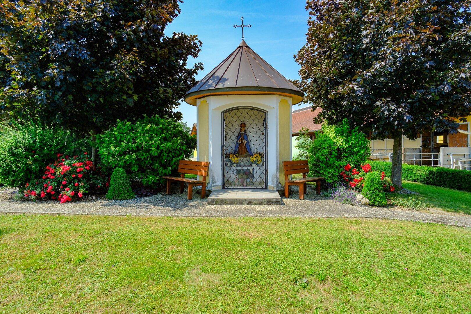 Kleine Kapelle in einem gepflegten Garten mit Bänken und Blumenbeeten.