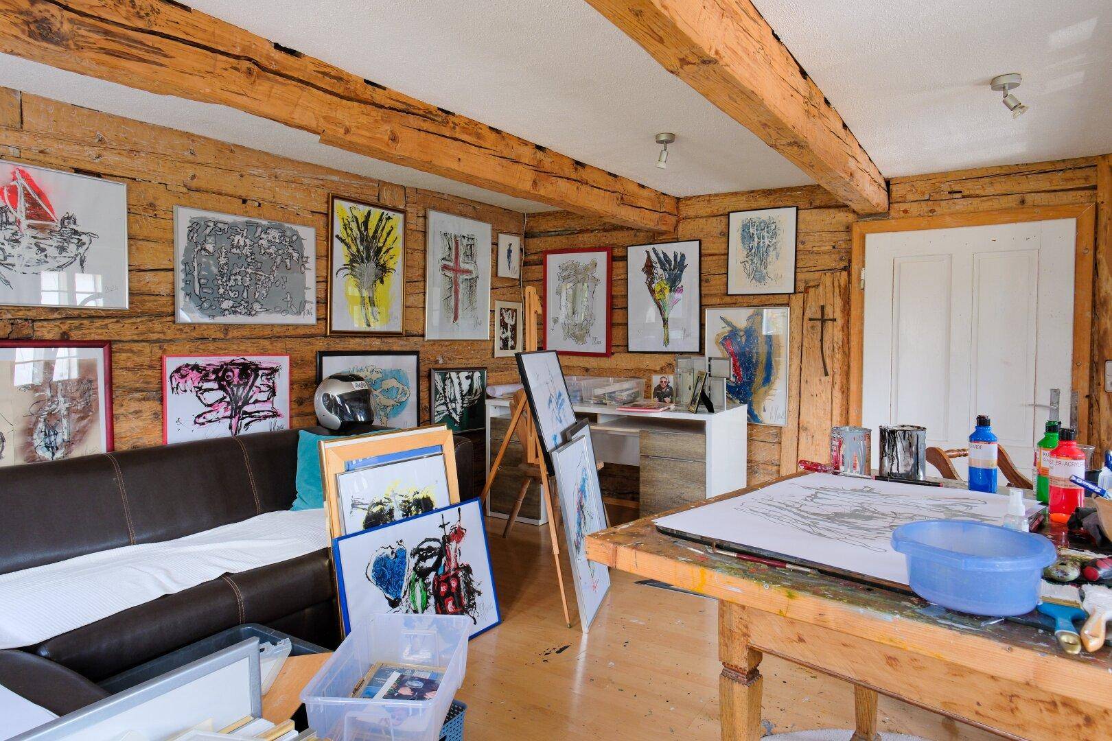 Künstleratelier mit Holzbalken, vielen Bildern und einem Arbeitstisch.