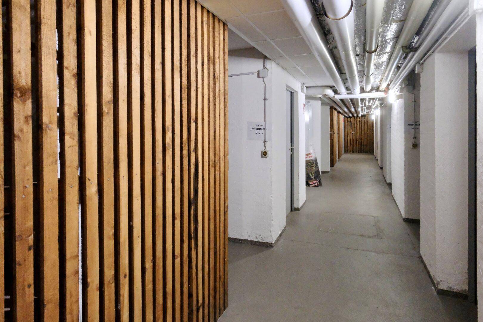 Langer Kellerflur mit Holzabteilen und sichtbaren Versorgungsleitungen an der Decke.