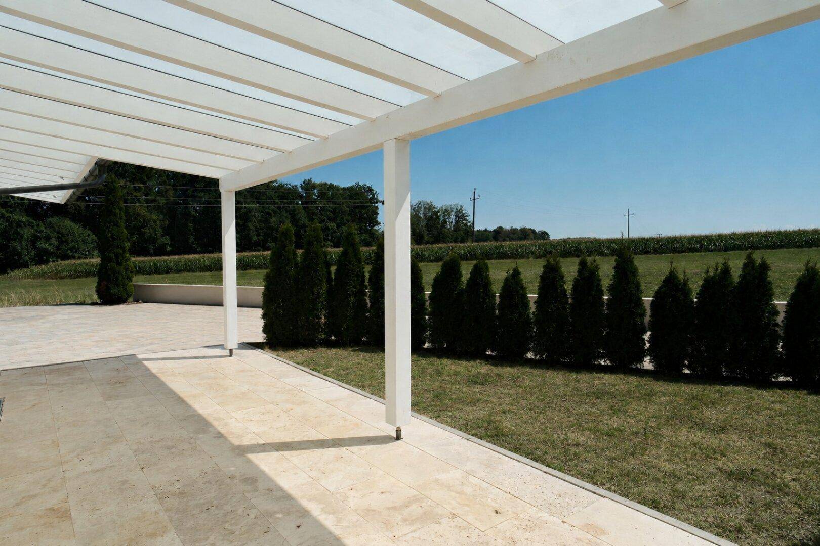 Helle Terrasse unter einer weißen Pergola mit Blick auf die grüne Landschaft.