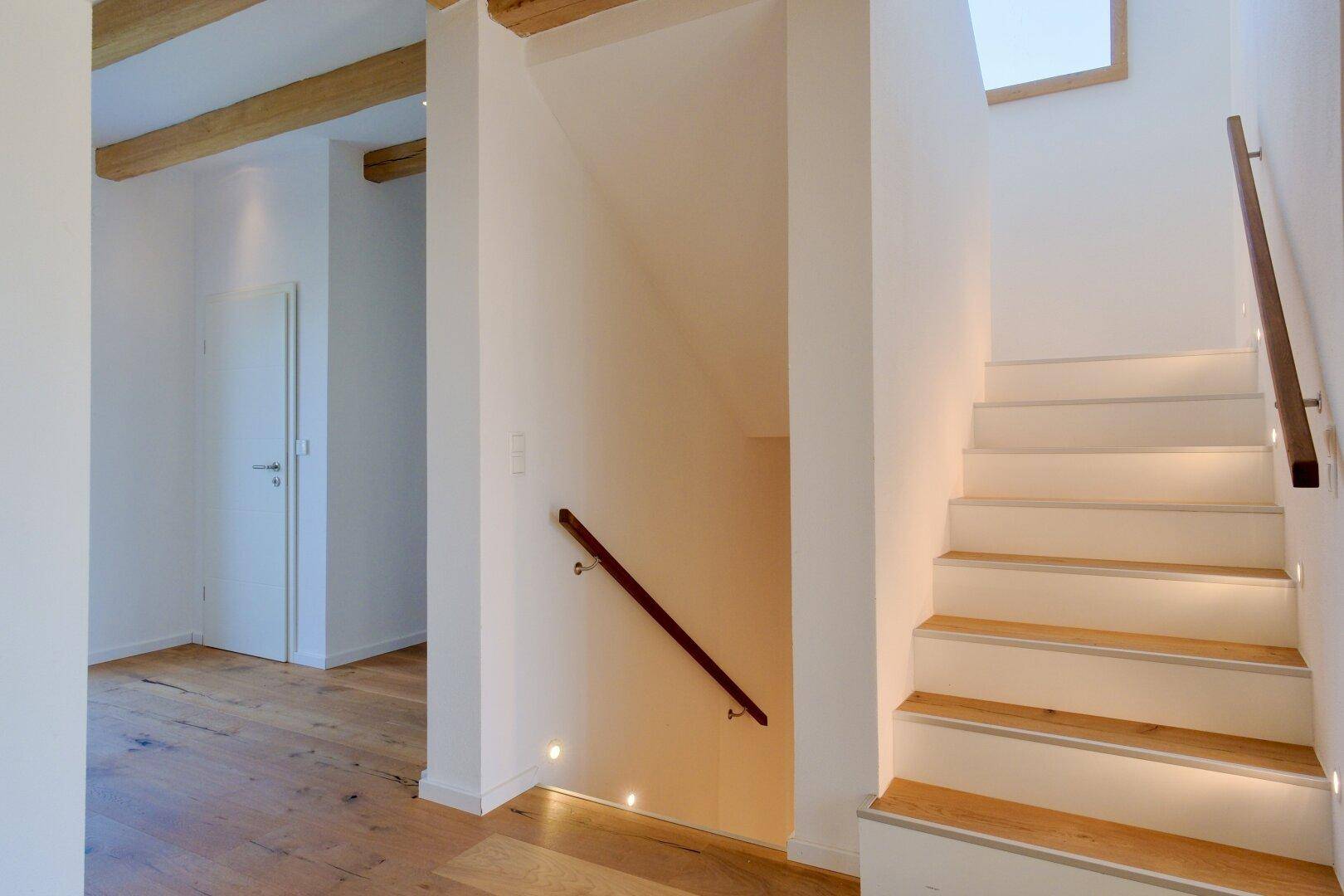 Heller Flur mit Holztreppe, integrierter Beleuchtung und sichtbaren Deckenbalken.