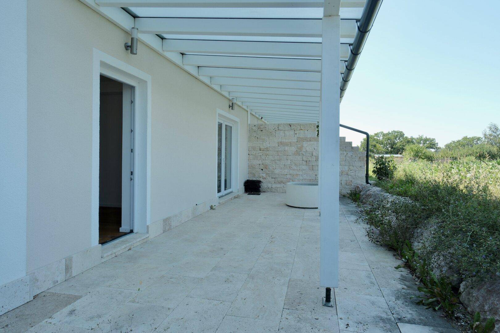 Überdachte Terrasse mit hellem Steinboden und Zugang zum Gartenbereich.