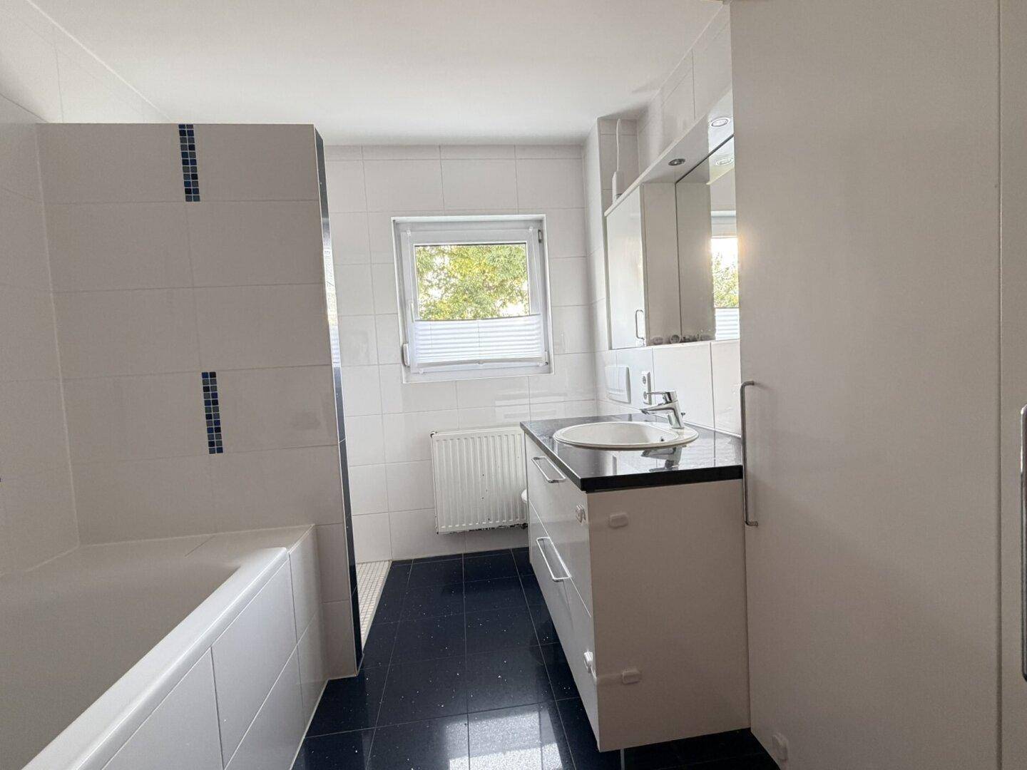 Helles Badezimmer mit weißen Wandfliesen, schwarzem Boden, Badewanne und Fenster.