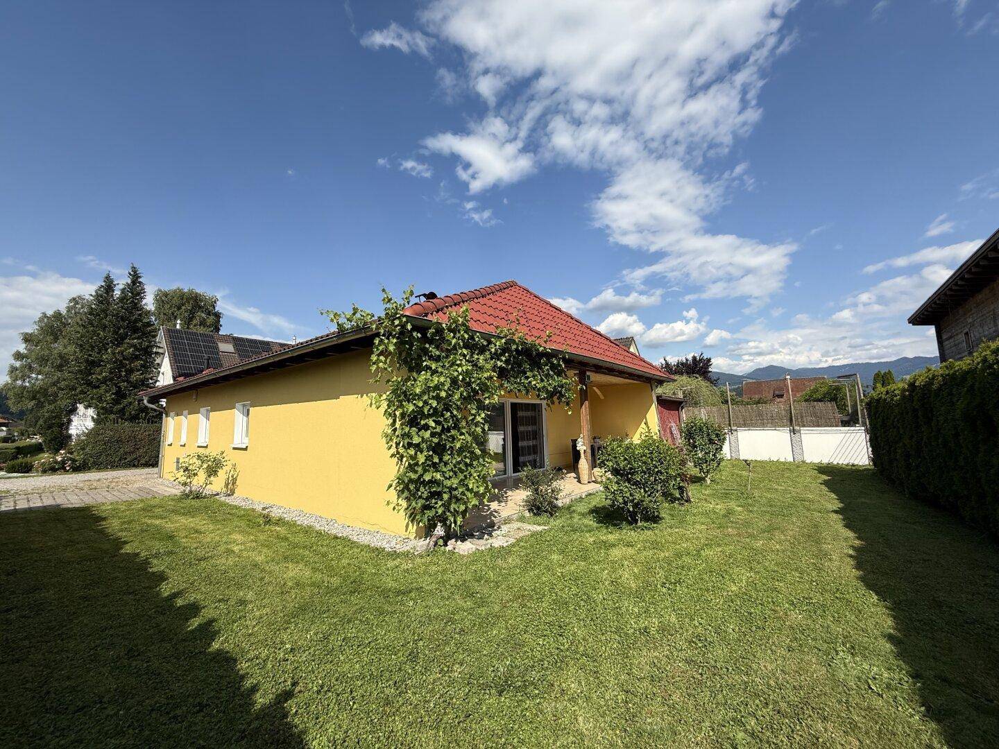 Gelbes Haus mit rotem Dach, umgeben von einem grünen Garten und Bäumen unter blauem Himmel.