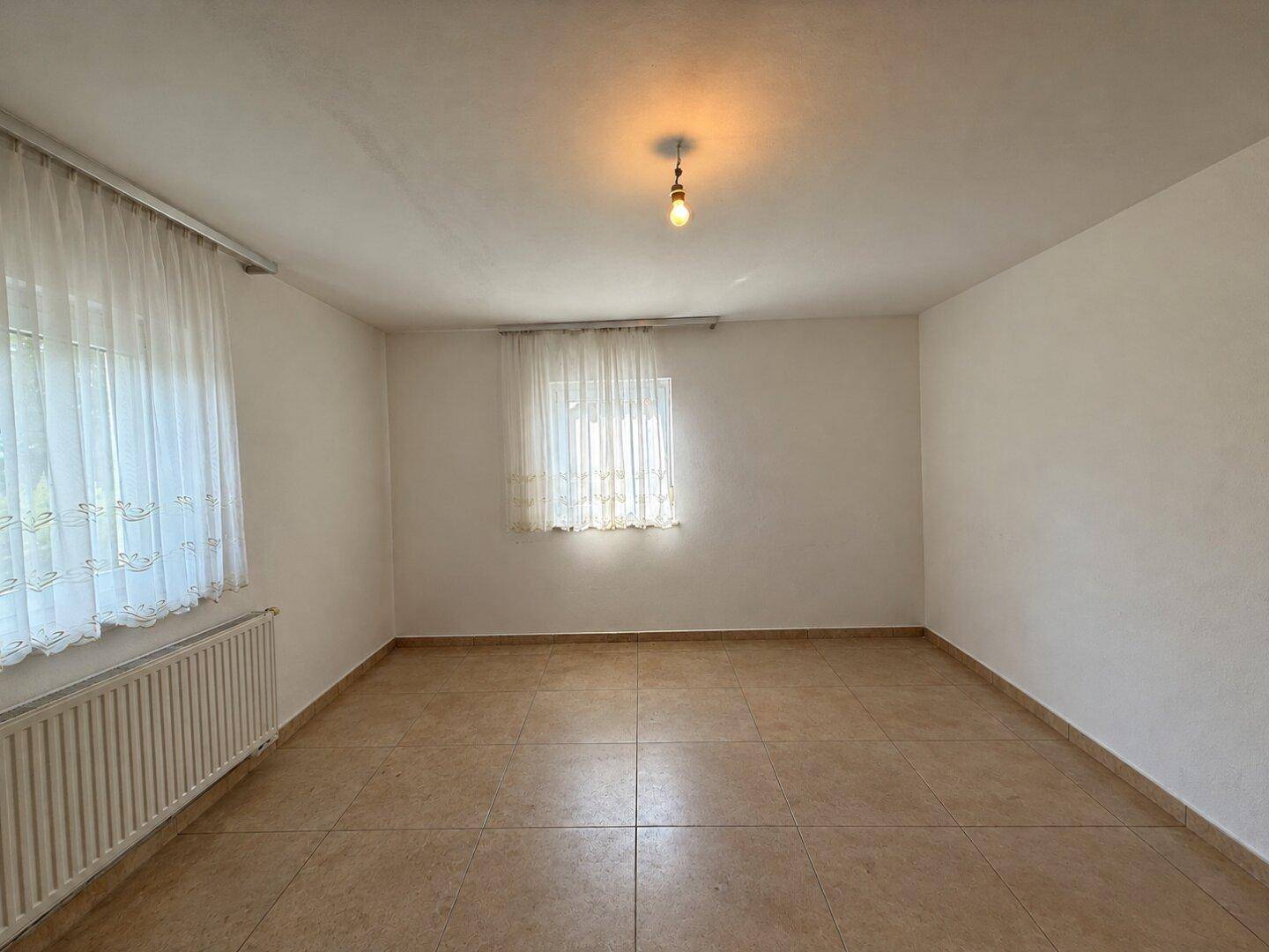 Helles Zimmer mit Fliesenboden, zwei Fenstern und Heizkörper, ideal als Schlaf- oder Arbeitszimmer.