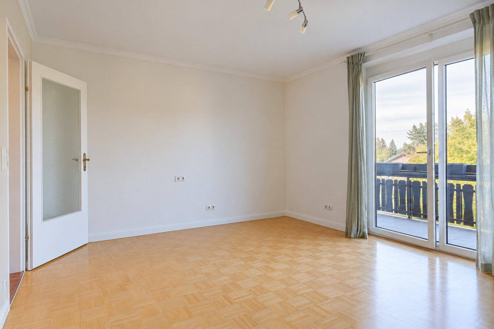 Helles, leeres Wohnzimmer mit Parkettboden und großem Fenster, das zum Balkon führt.