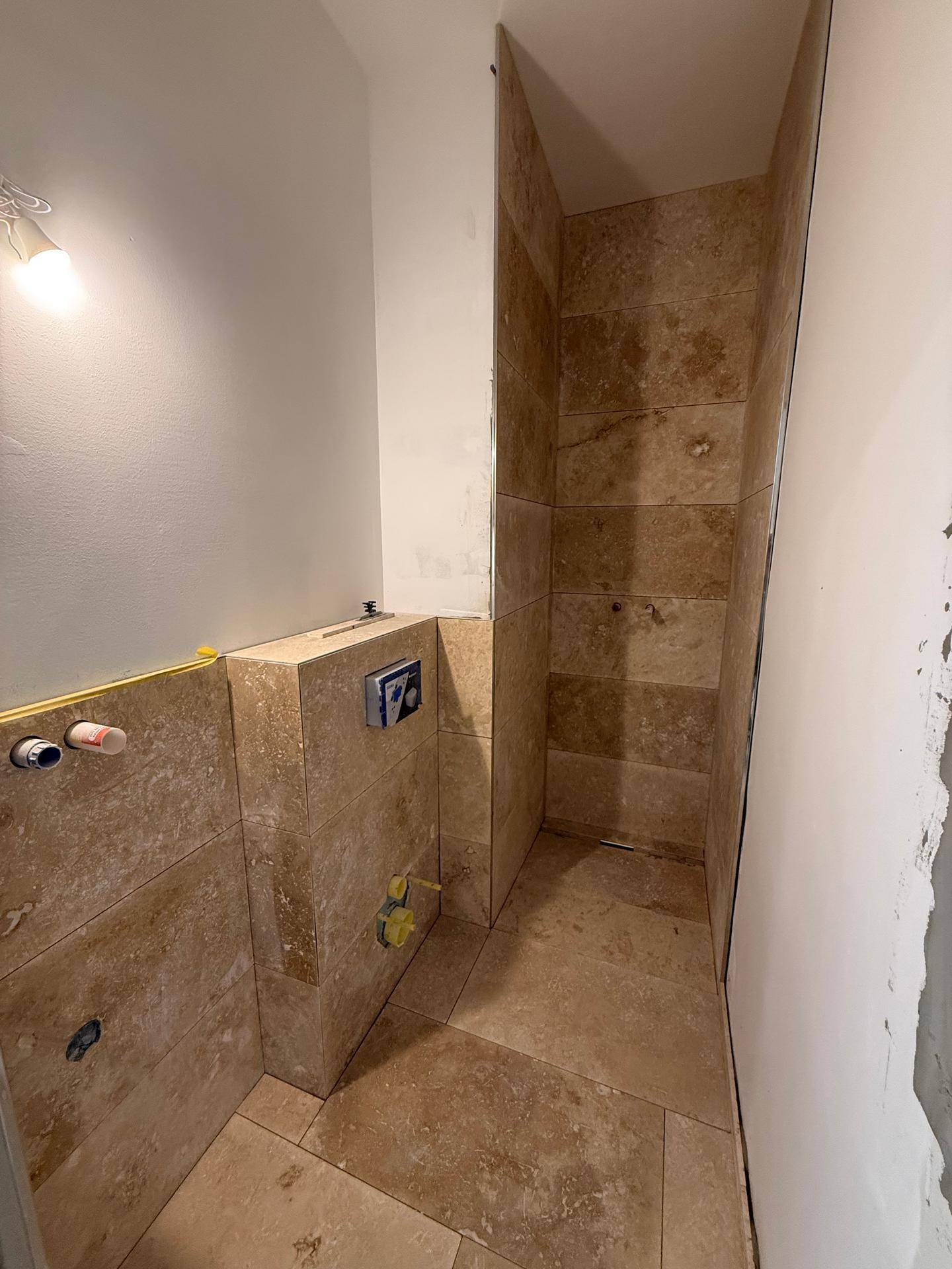 Badezimmer im Bau mit beigefarbenen Natursteinfliesen und vorbereiteten Installationen für die Dusche.