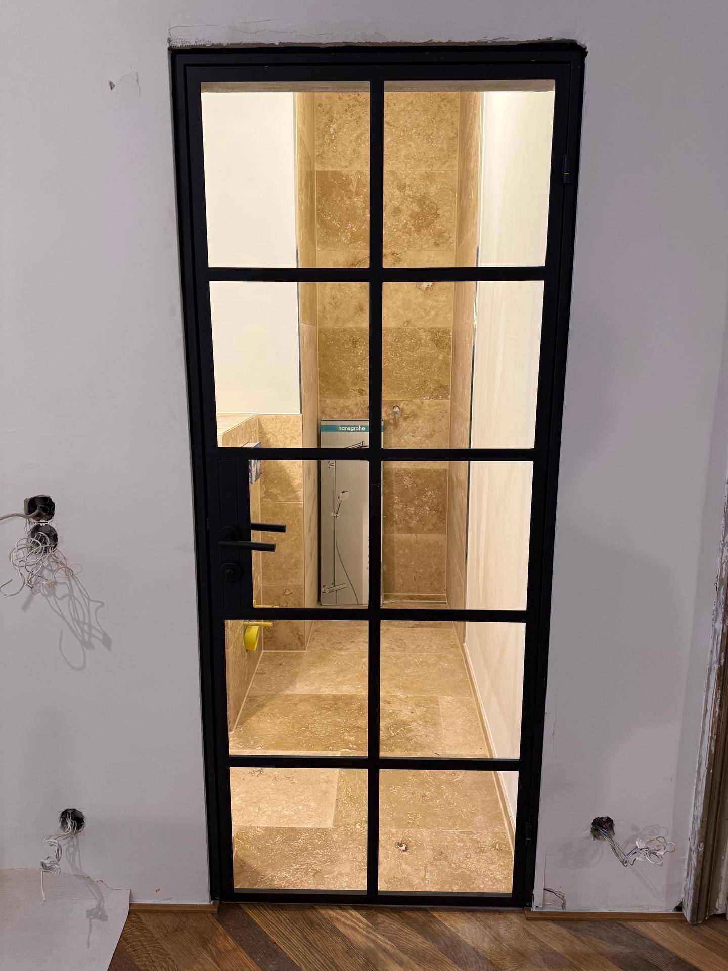 Badezimmer im Bau, sichtbar durch eine moderne schwarze Glastür, mit Natursteinfliesen.