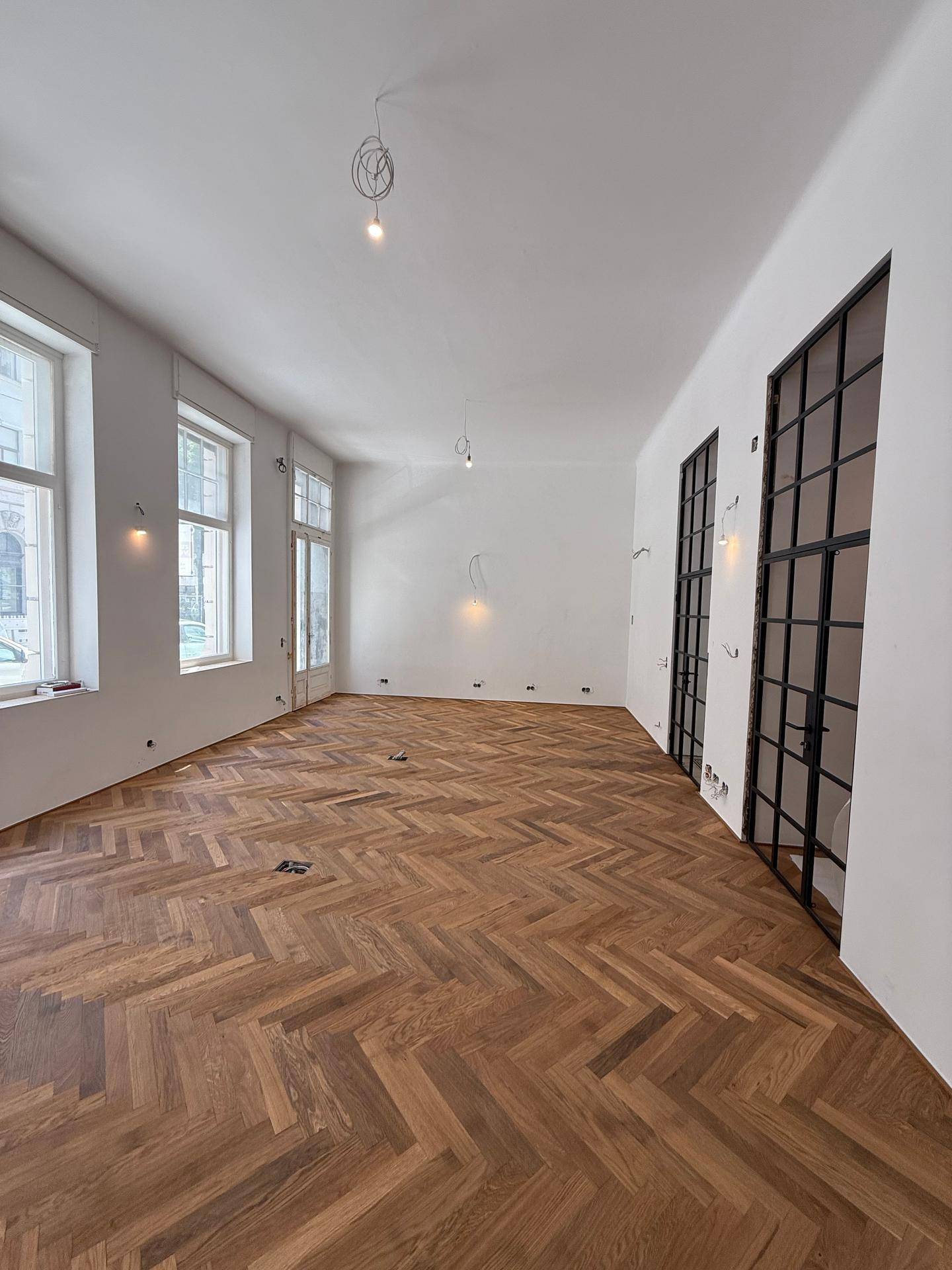 Heller, leerer Raum mit Fischgrätparkett, hohen Fenstern und modernen Glastüren.