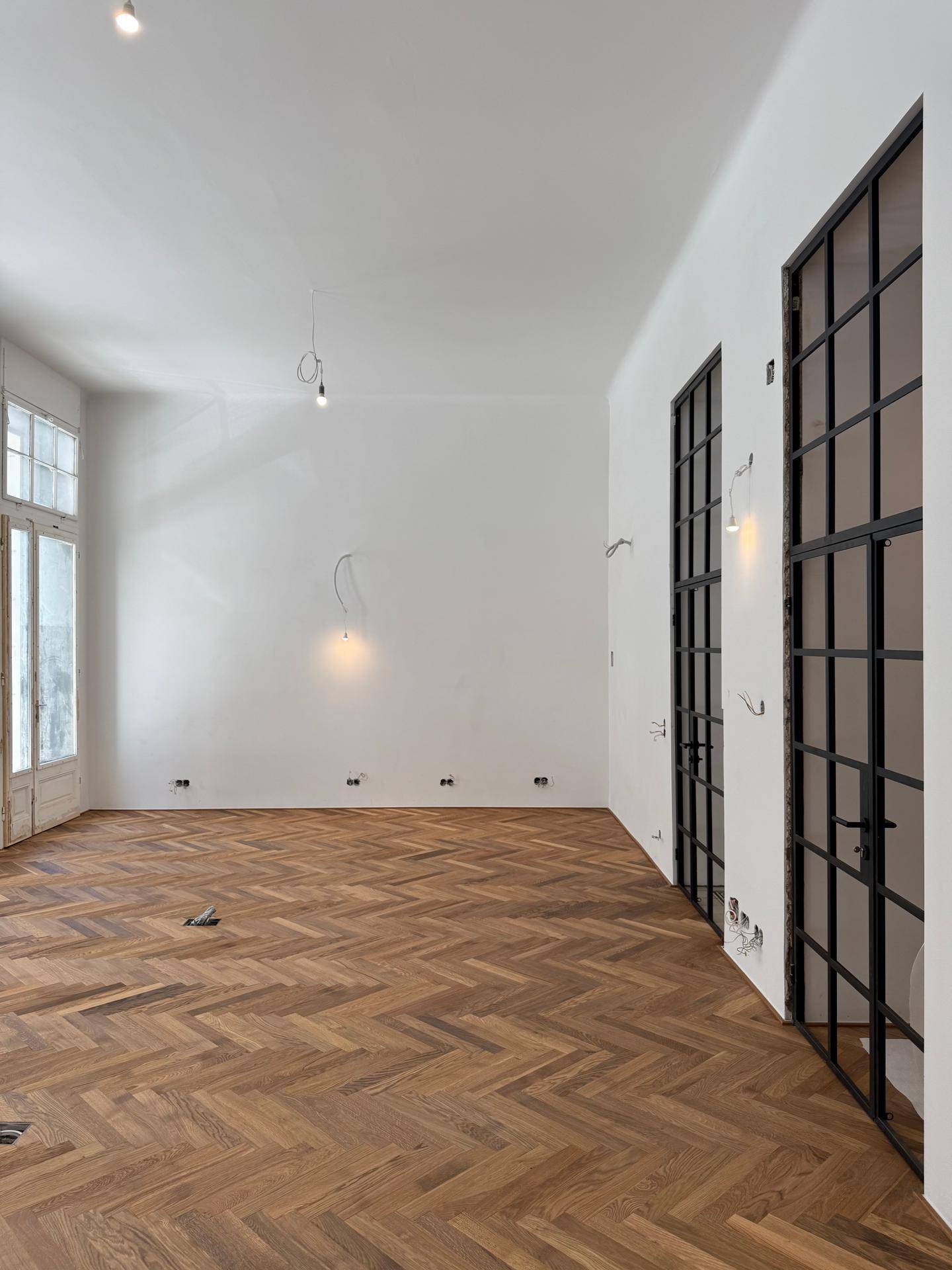 Heller, leerer Raum mit Fischgrätparkett, hohen Fenstern und modernen Glastüren.