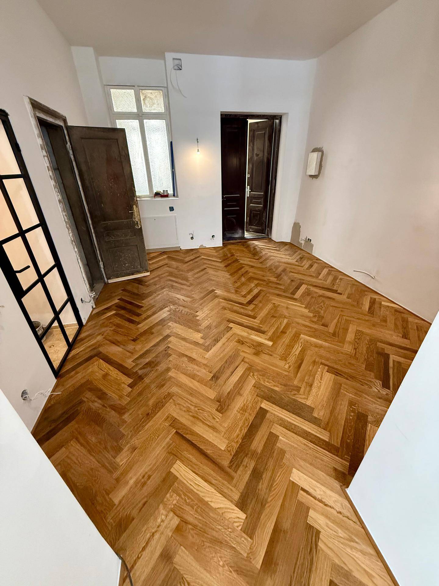 Raum mit Fischgrätparkett, hohen Decken und einer alten Holztür, die geöffnet ist.
