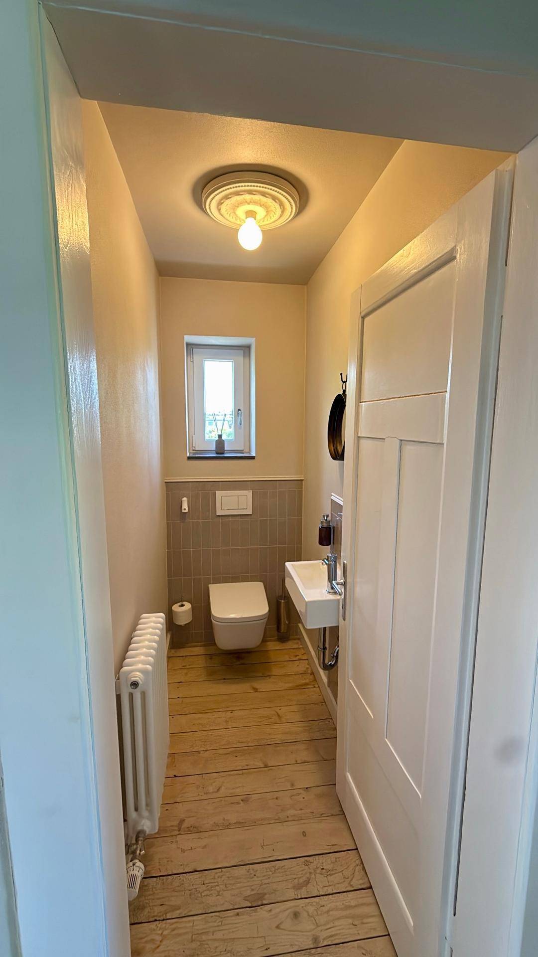 Schmaler WC-Raum mit Fenster, Holzfußboden, Heizkörper und modernem Waschbecken.