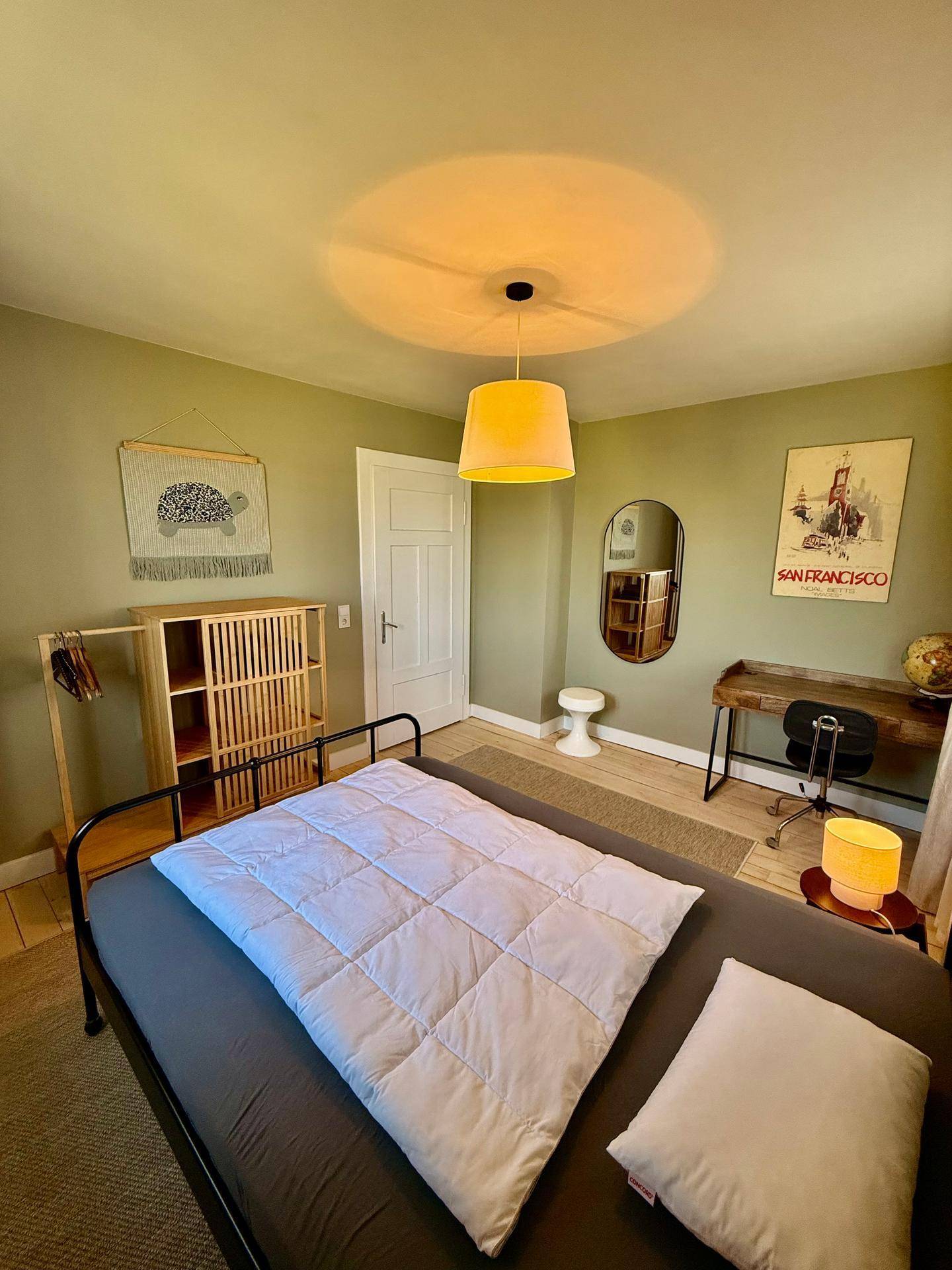 Helles Schlafzimmer mit olivgrünen Wänden, einem Metallbett und einem Schreibtisch.