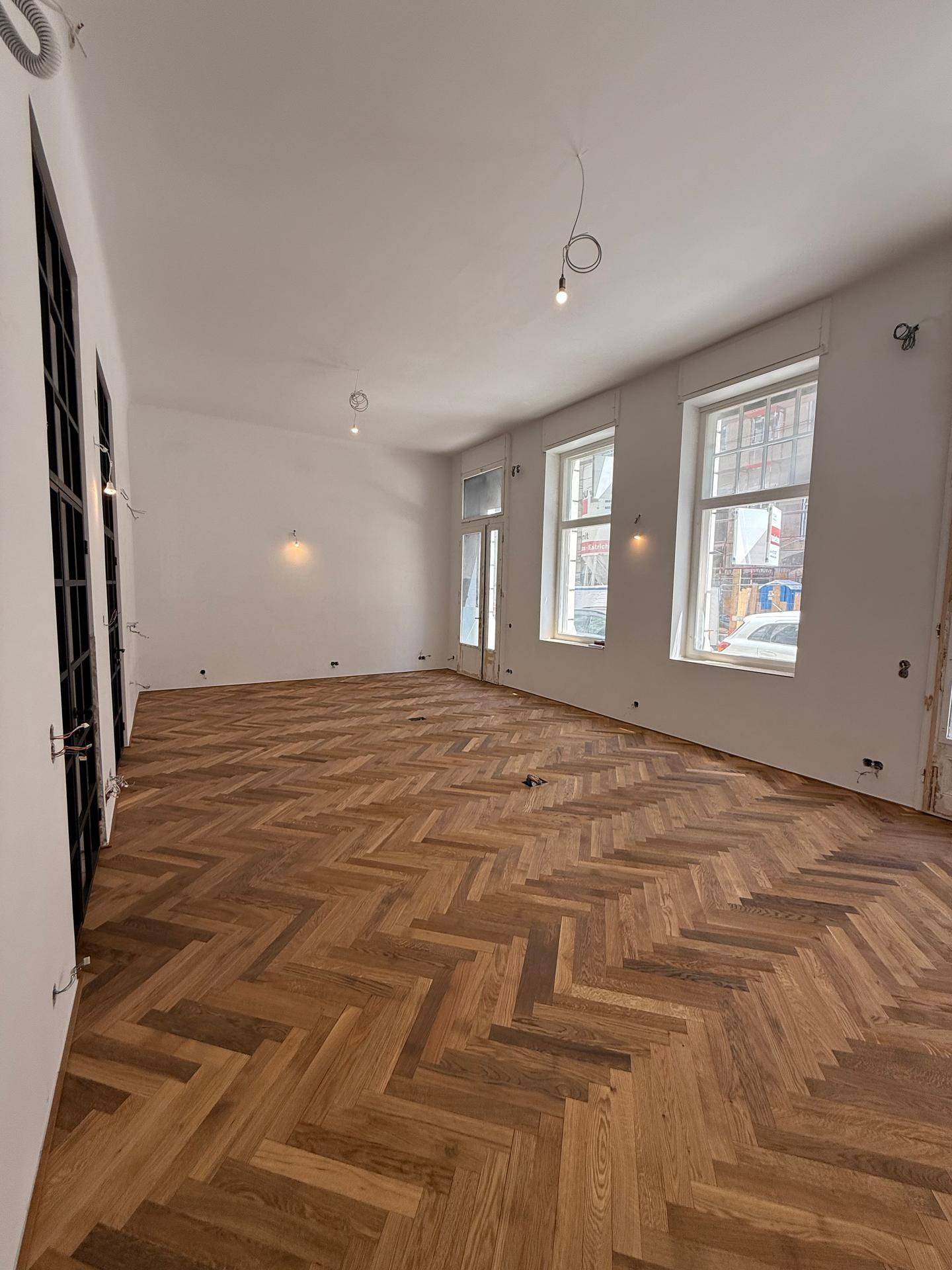 Heller Raum mit Fischgrätparkett, großen Fenstern zur Straße und modernen Glastüren.