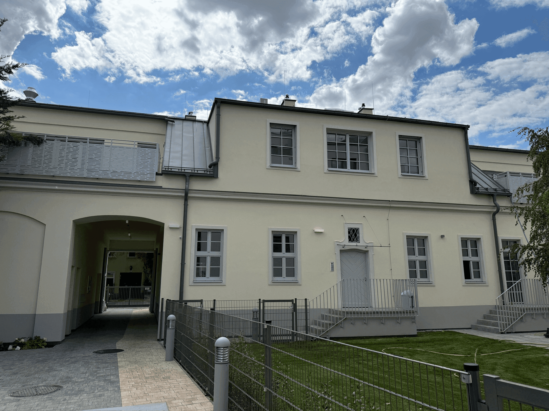 Rückseite eines Gebäudes mit hellem Anstrich, grauen Fenstern, einem Torbogen zum Innenhof und einem kleinen Rasenbereich.