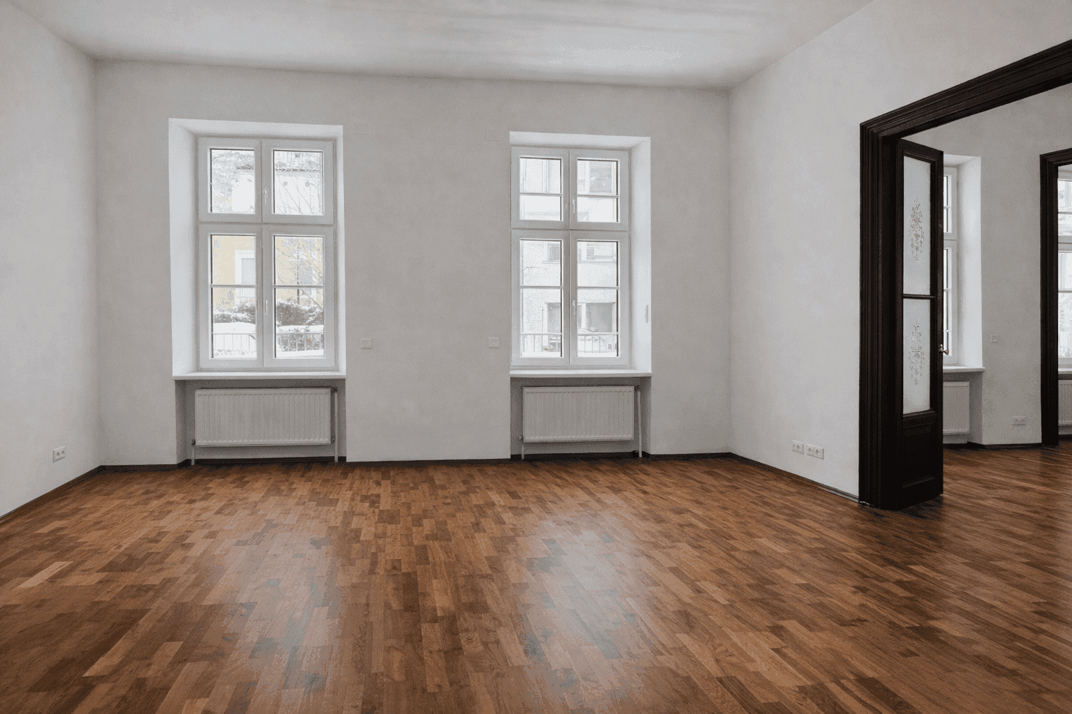 Helles Wohnzimmer mit klassischem Parkettboden, zwei großen Fenstern und einer dunklen Flügeltür, die in einen Nebenraum führt.