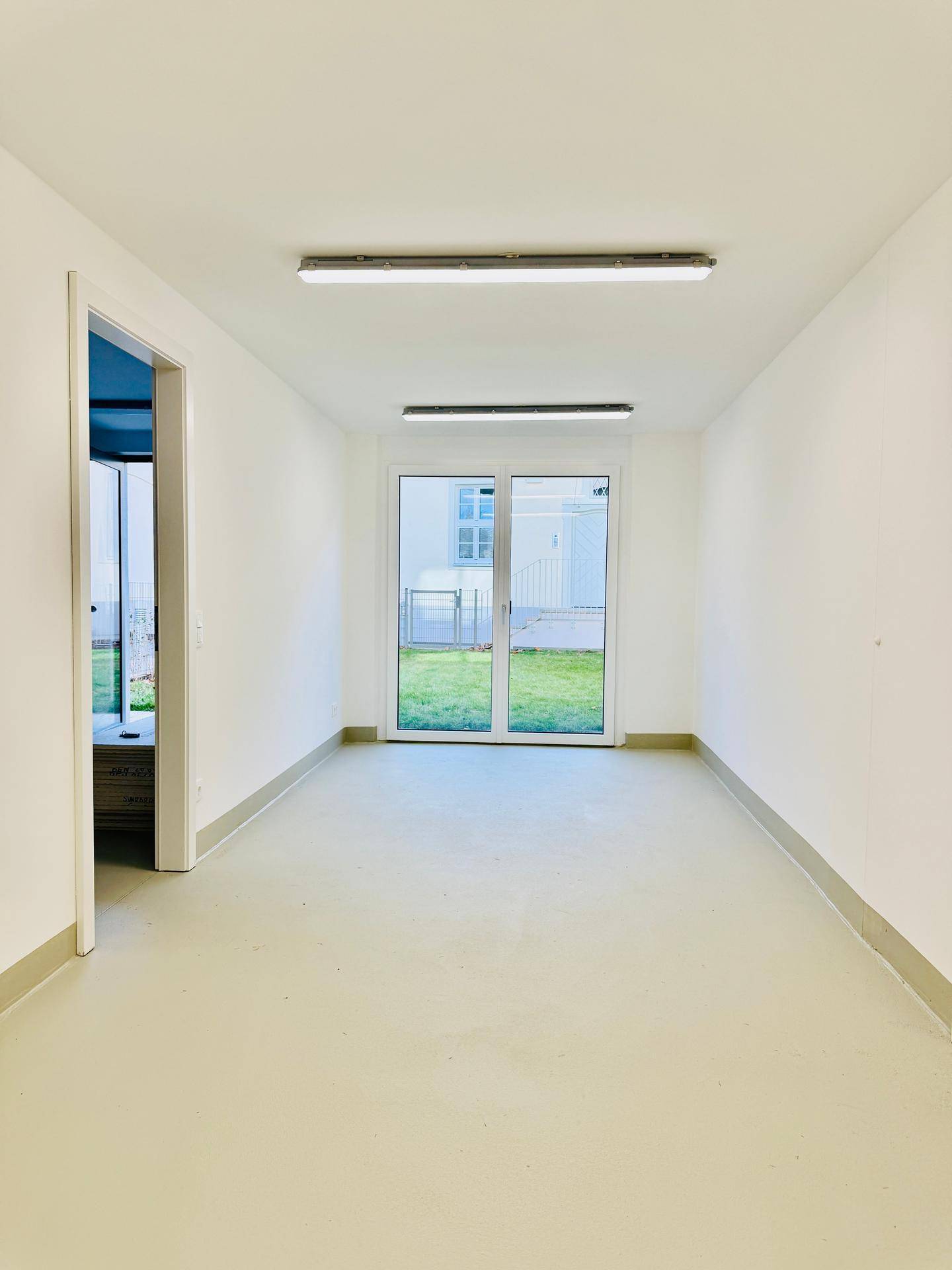 Heller, leerer Raum mit hellem Boden, weißen Wänden und einer Glastür, die direkt zum Rasen führt.