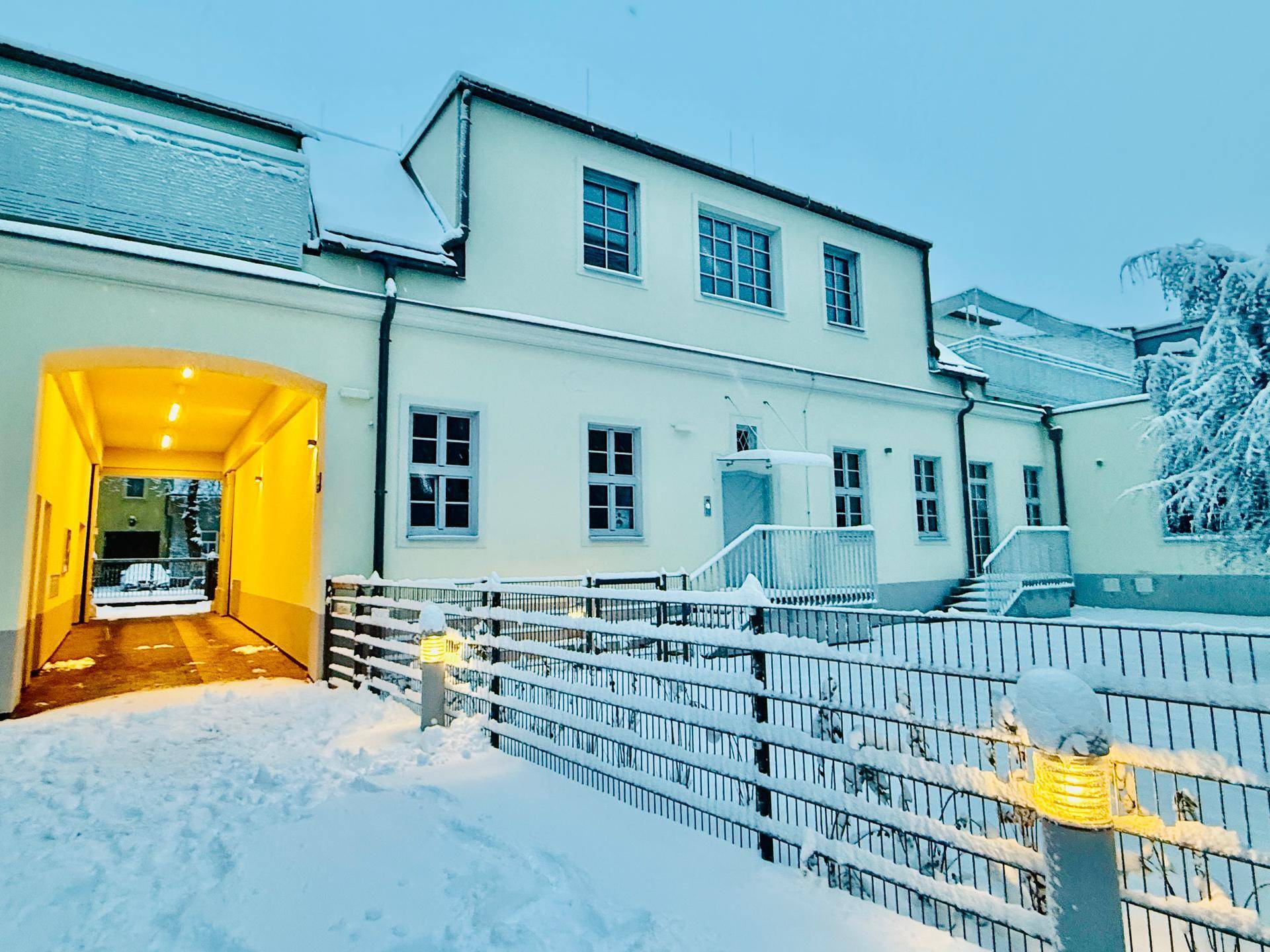 Winterliche Szene des Gebäudes mit Schnee, einem hell erleuchteten Torbogen zum Innenhof und blauen Fenstern.