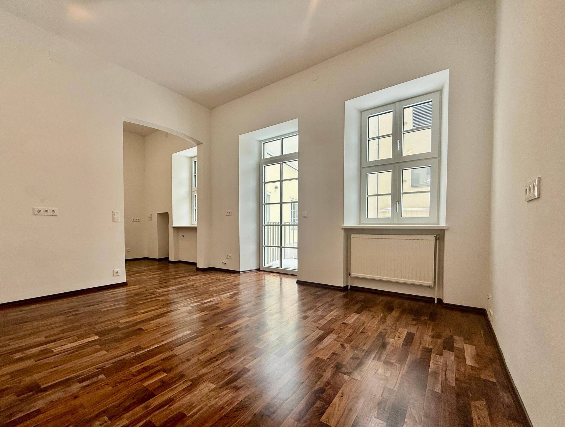 Heller Wohnraum mit Holzboden, hohen Decken und einer Fenstertür, die zu einem französischen Balkon führt.