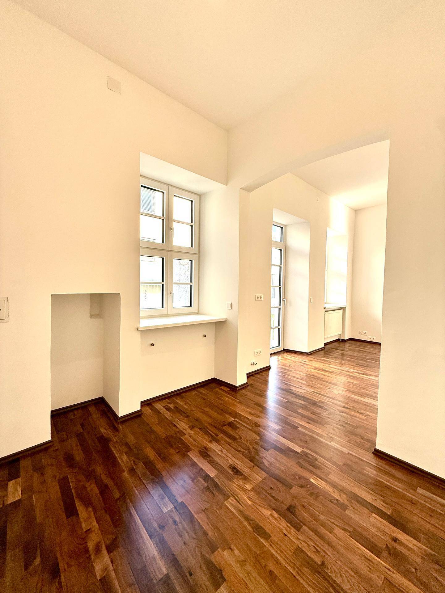 Geräumiger Wohnbereich mit glänzendem Parkettboden, hohen Fenstern und einem offenen Durchgang zu einem weiteren Zimmer.