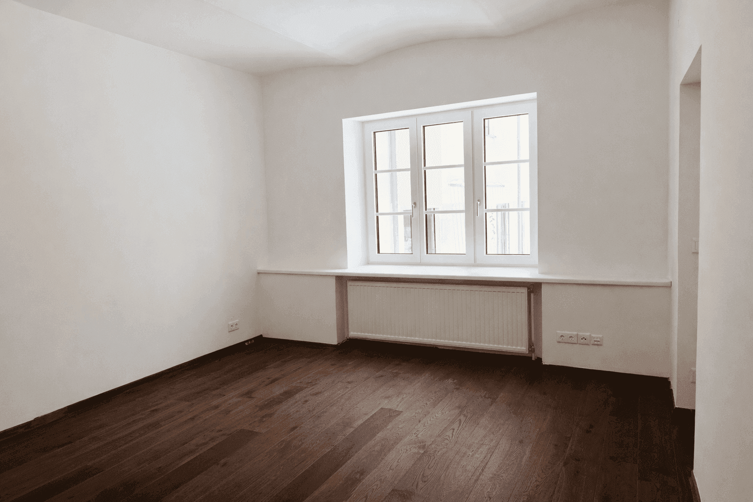 Ruhiges Schlafzimmer mit dunklem Holzboden, weißen Wänden und einem großen Fenster mit Heizkörper darunter.